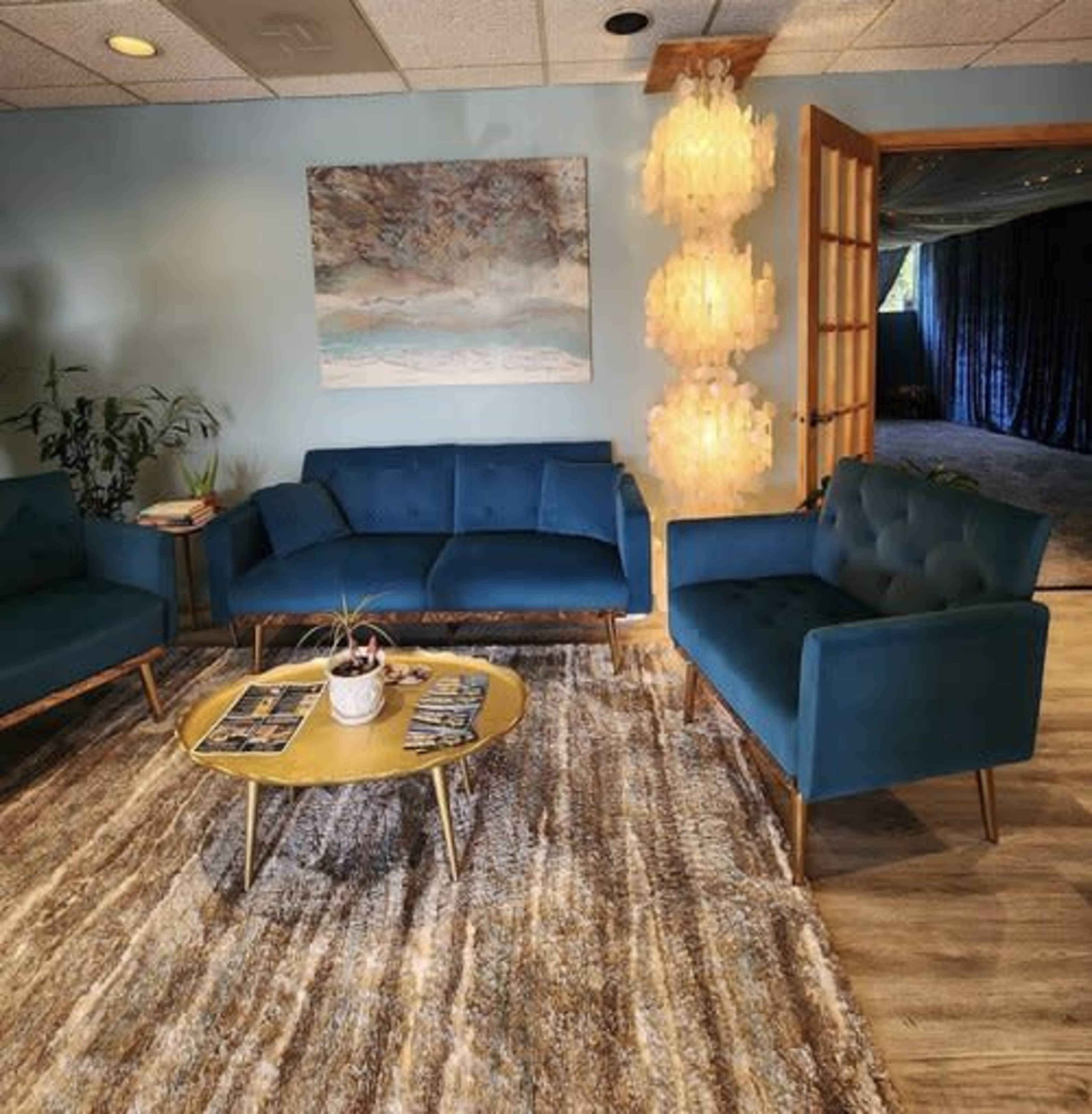 A modern seating area with teal sofas, a round coffee table, and a decorative light fixture, set against a textured rug and light blue walls.