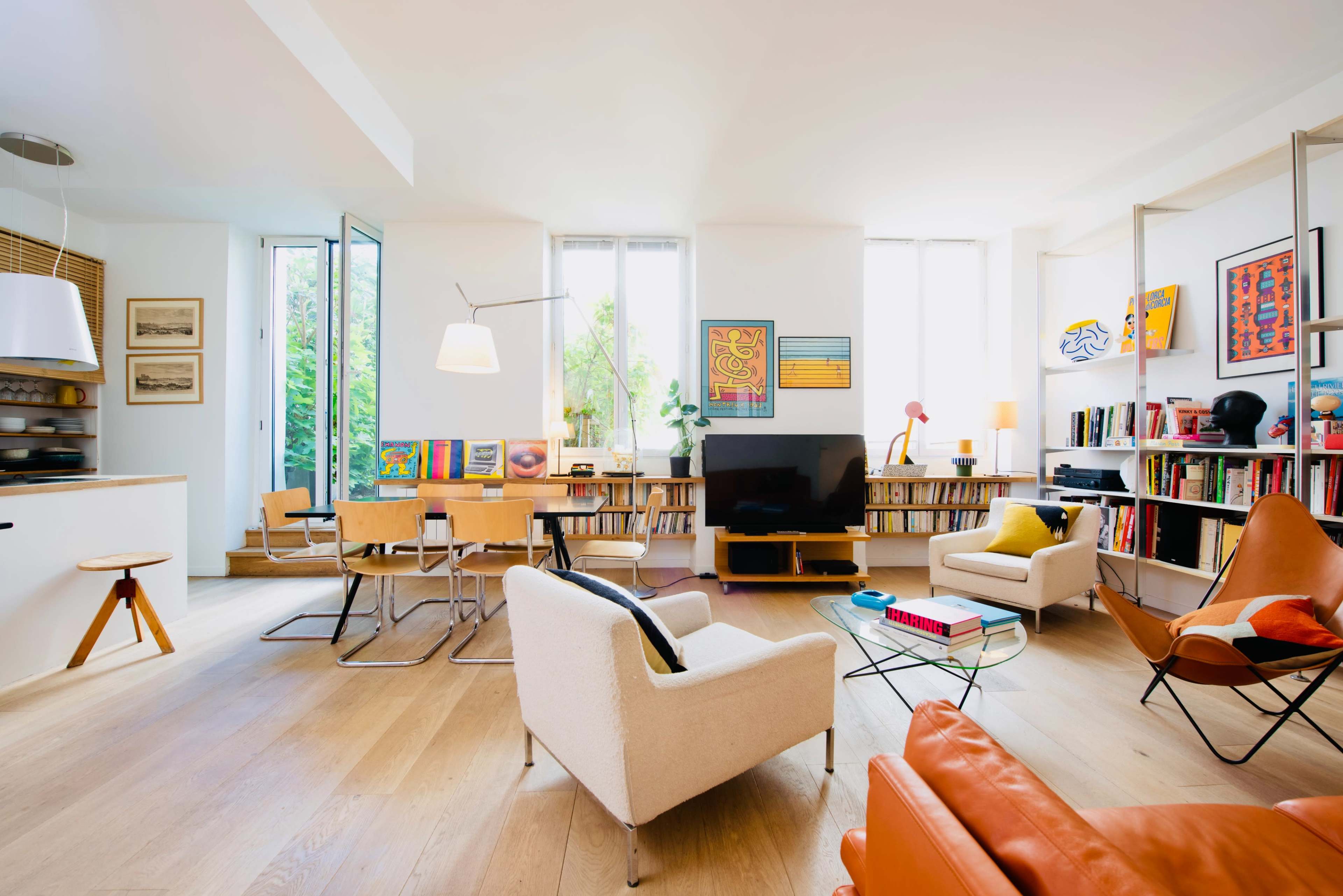 A modern living room with wooden flooring, a television, bookshelves filled with books, and various seating arrangements, including a white armchair and an orange leather chair.