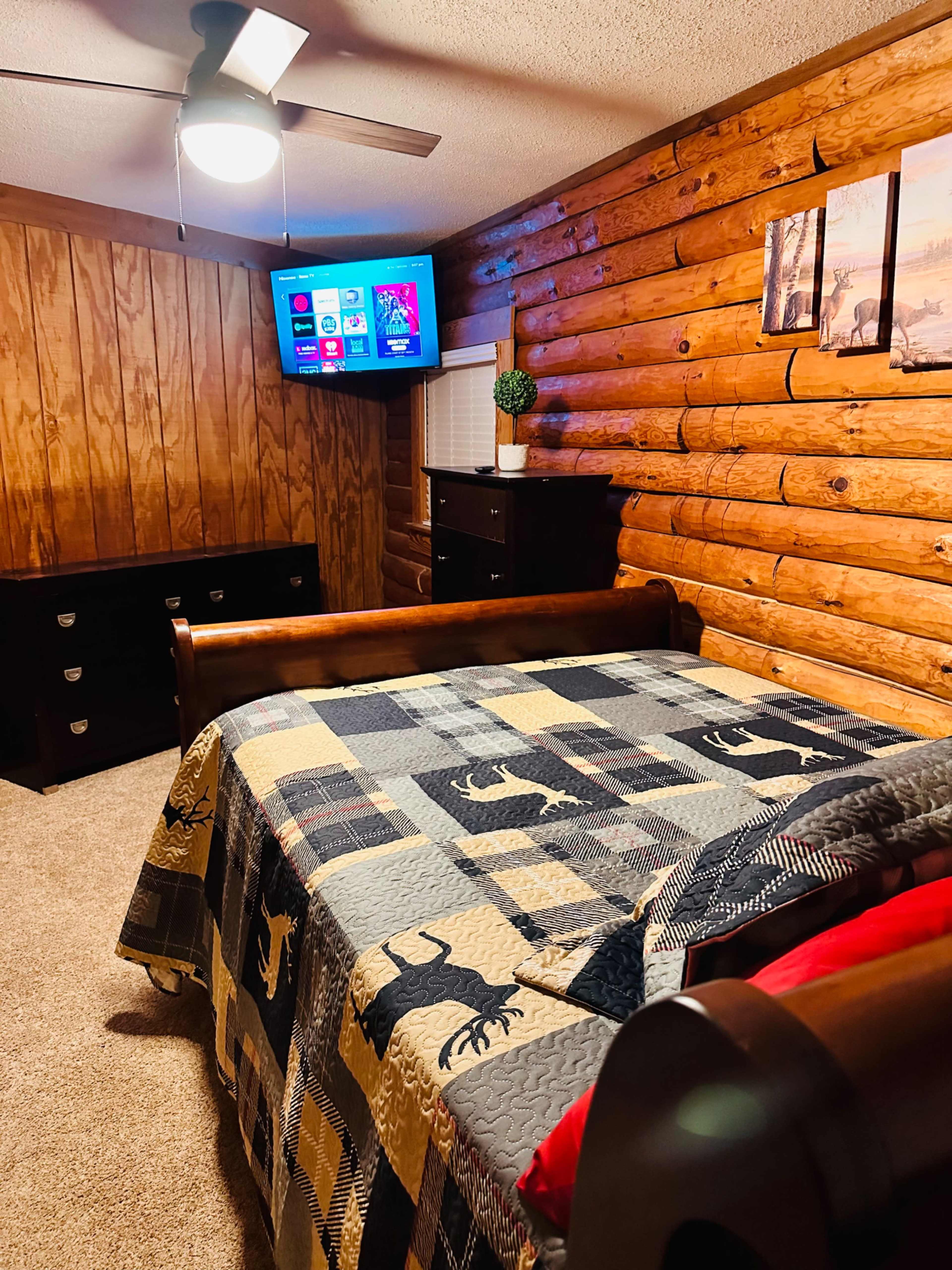 A cozy bedroom featuring a wooden log wall, a bed with a patterned quilt, a television mounted on the wall, and a dresser.
