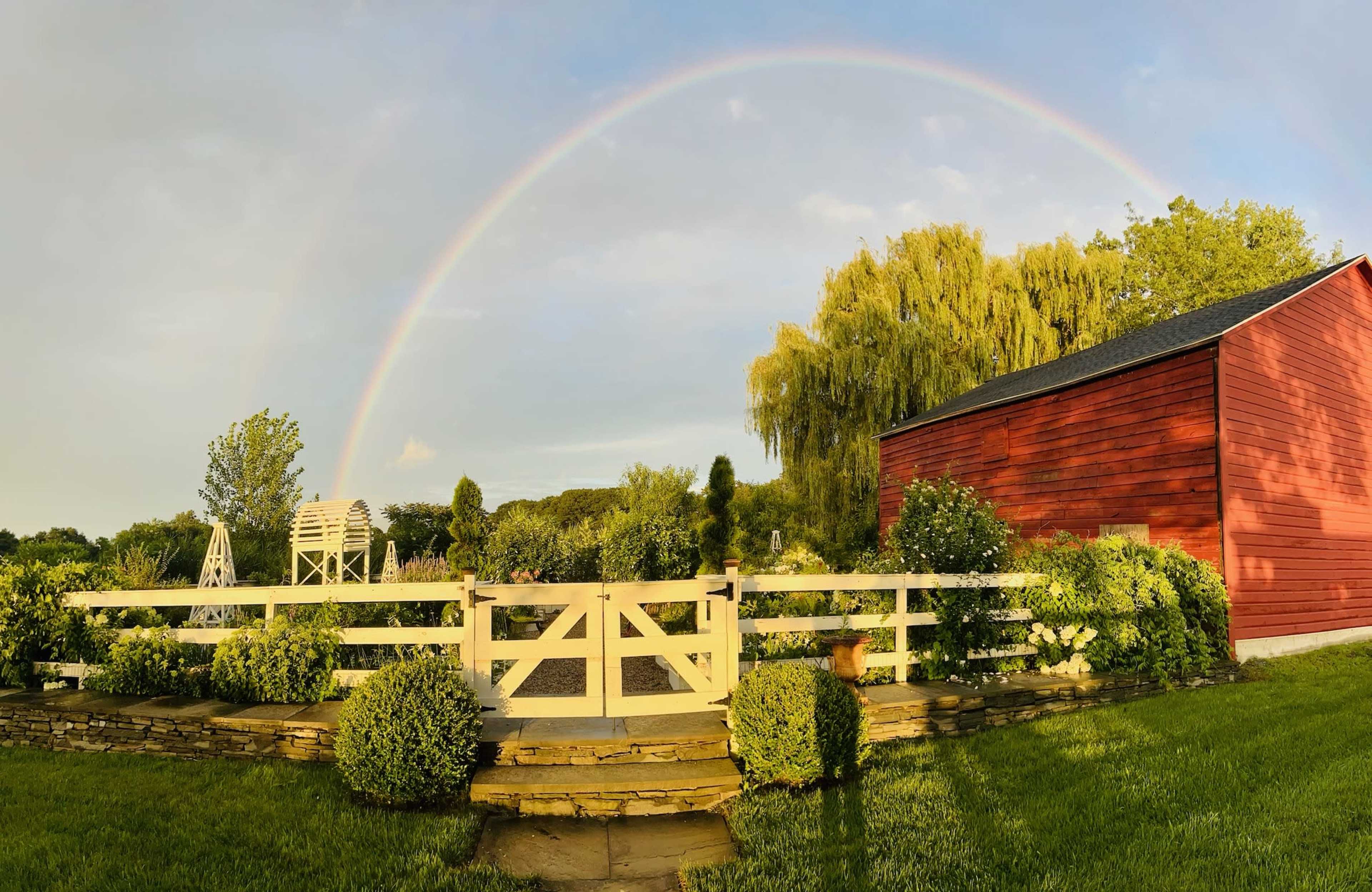 A vibrant rainbow arcs over a red barn surrounded by lush greenery and neatly trimmed hedges.