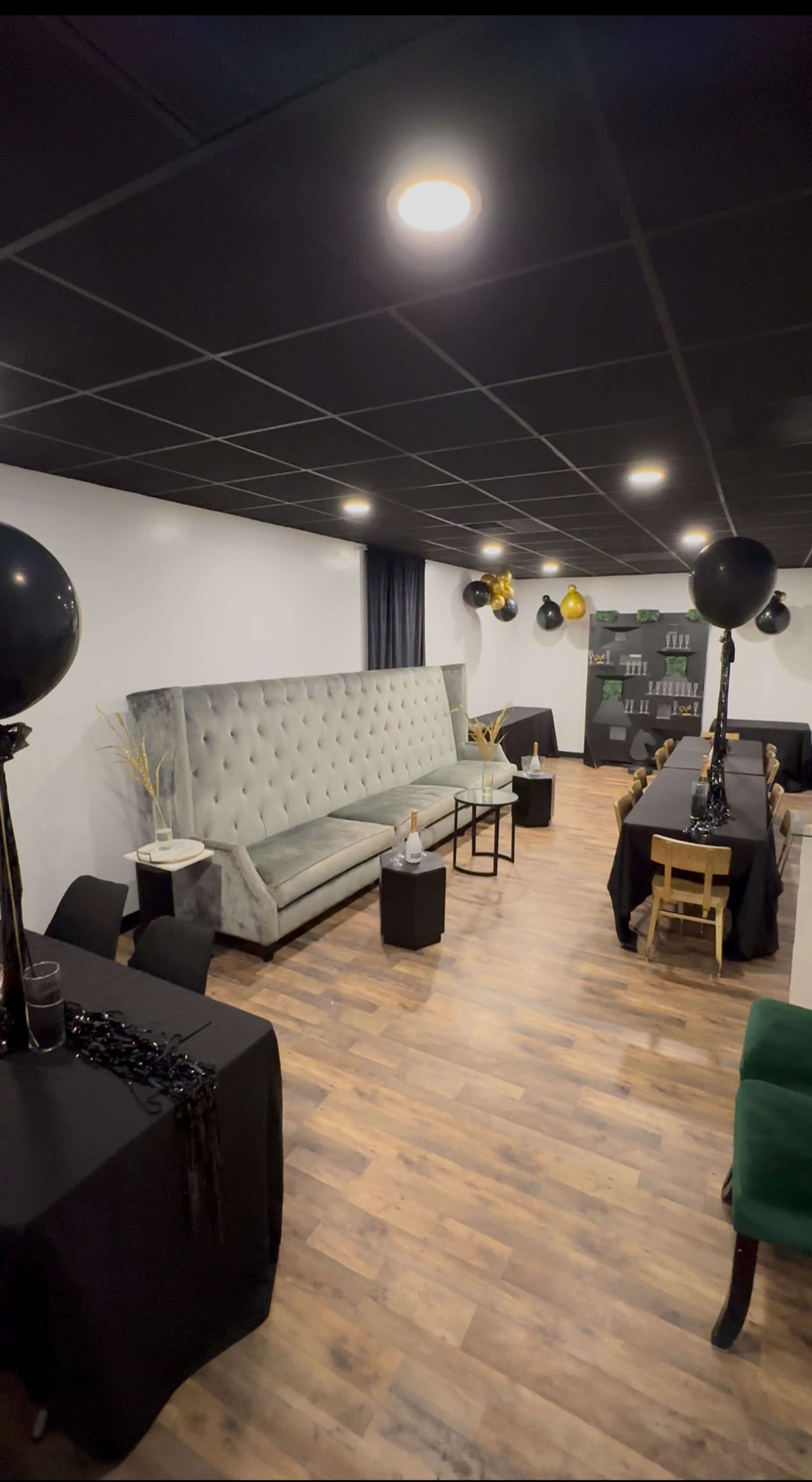 The image shows a modern event space with gray tufted seating, black tables, and festive decorations including black and gold balloons.
