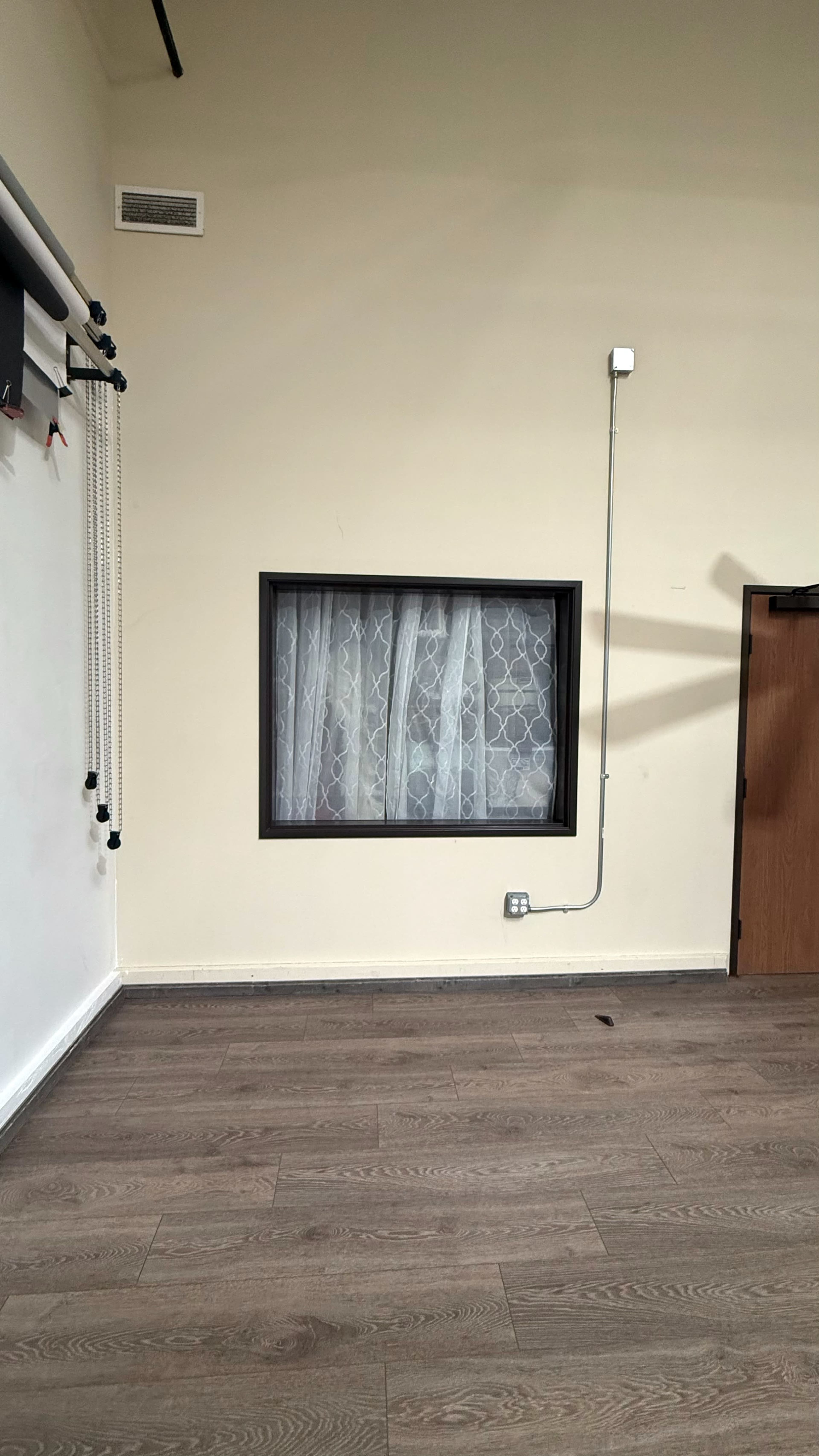 The image shows a room with a window featuring sheer curtains, a wooden door on one side, and a wall-mounted cable running vertically next to the window.