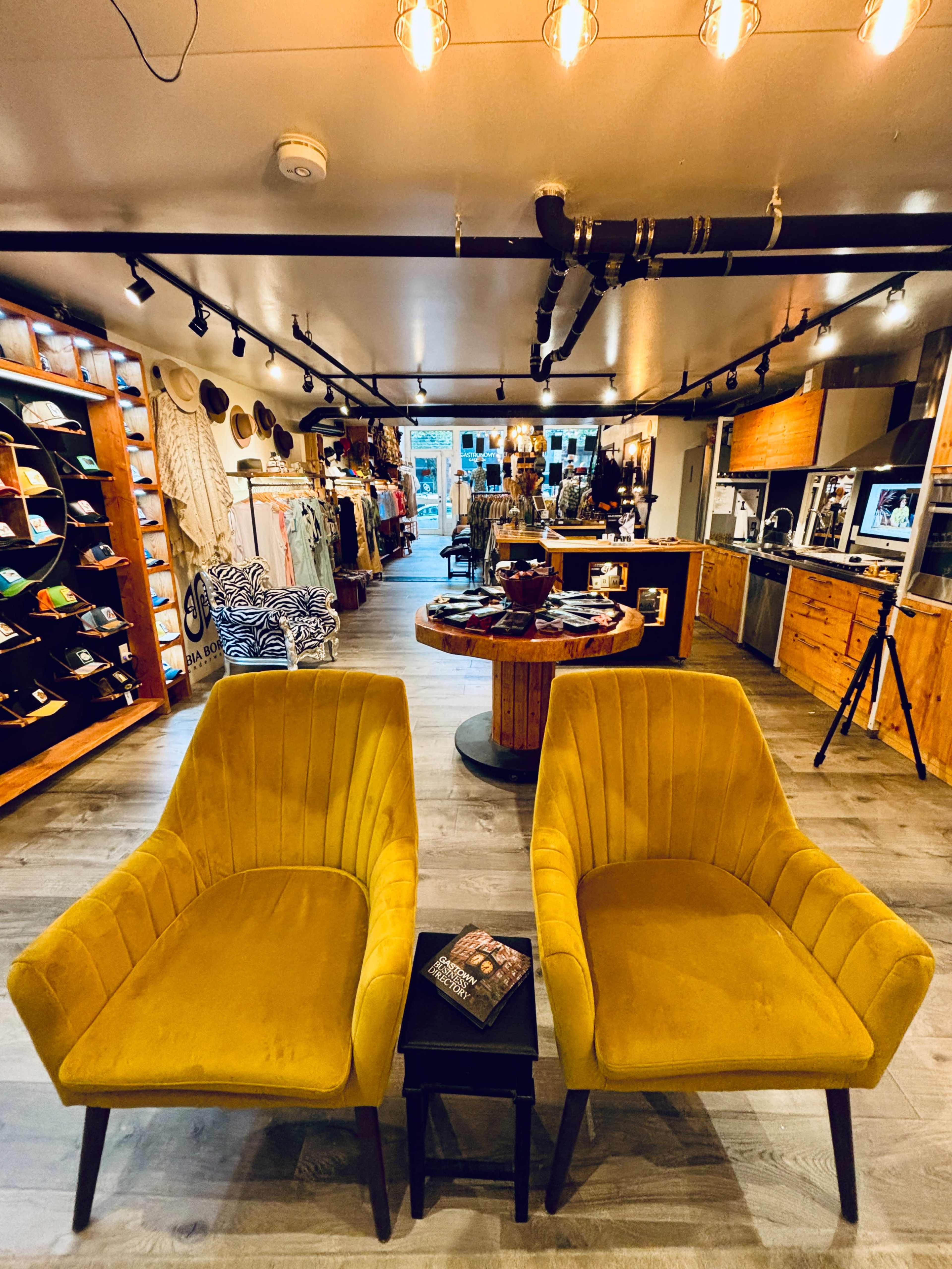 A boutique interior featuring two mustard-colored armchairs beside a small black table, with various clothing and accessories displayed on shelves in the background.
