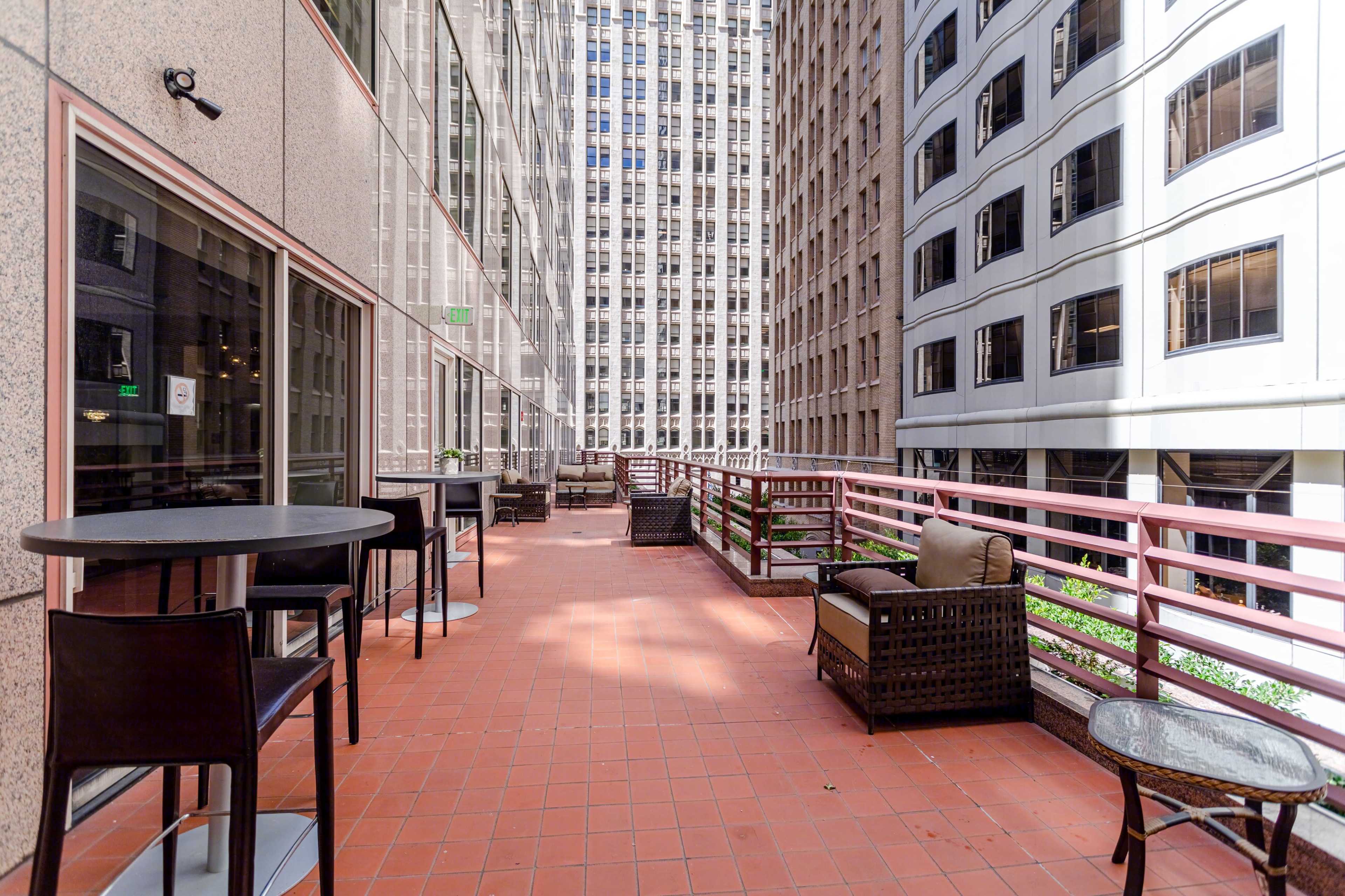 A balcony area with several tables and chairs overlooking a narrow atrium between tall buildings.