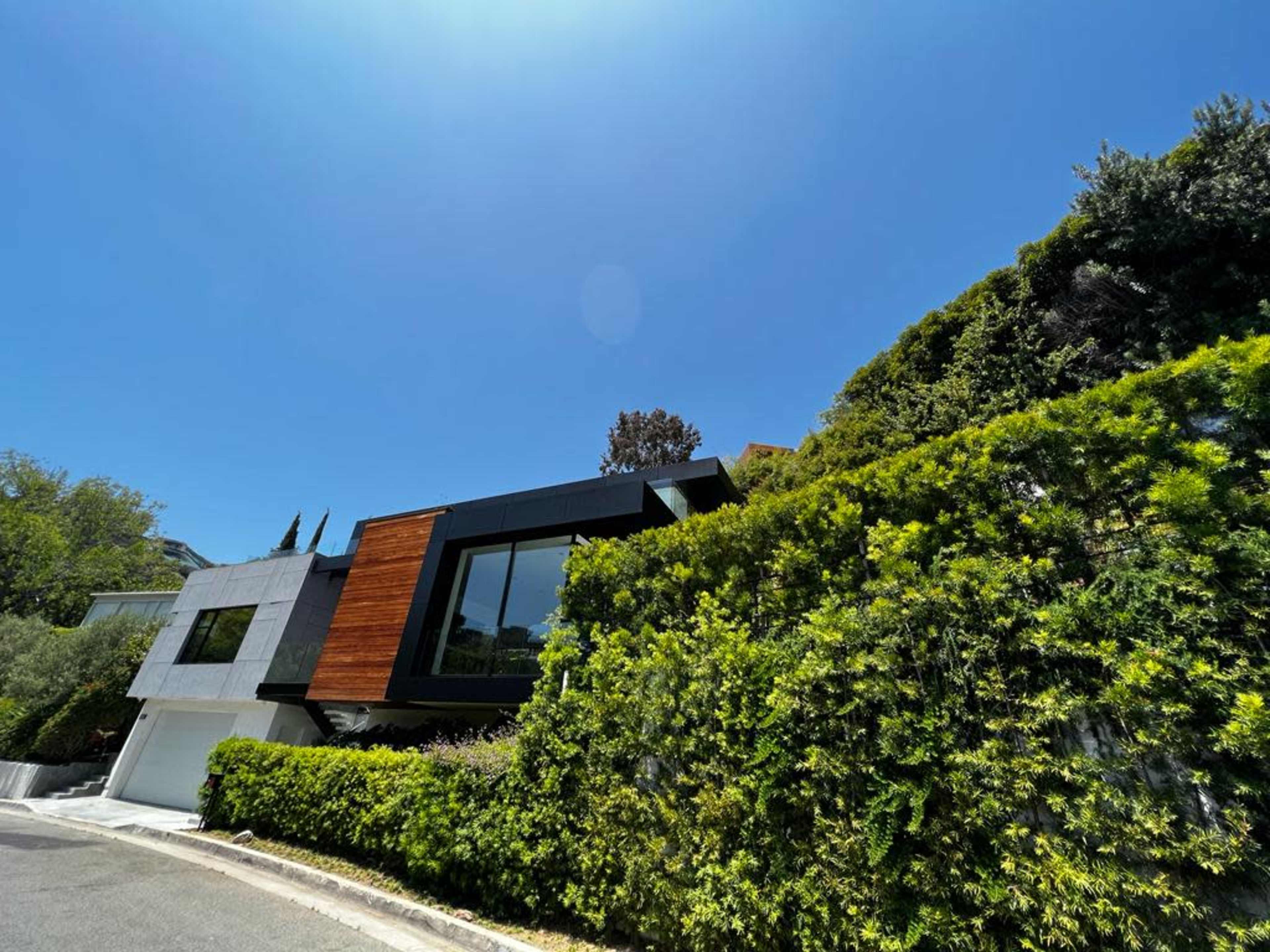 A modern house with a mix of wood and concrete siding is set on a hillside, surrounded by lush greenery under a clear blue sky.