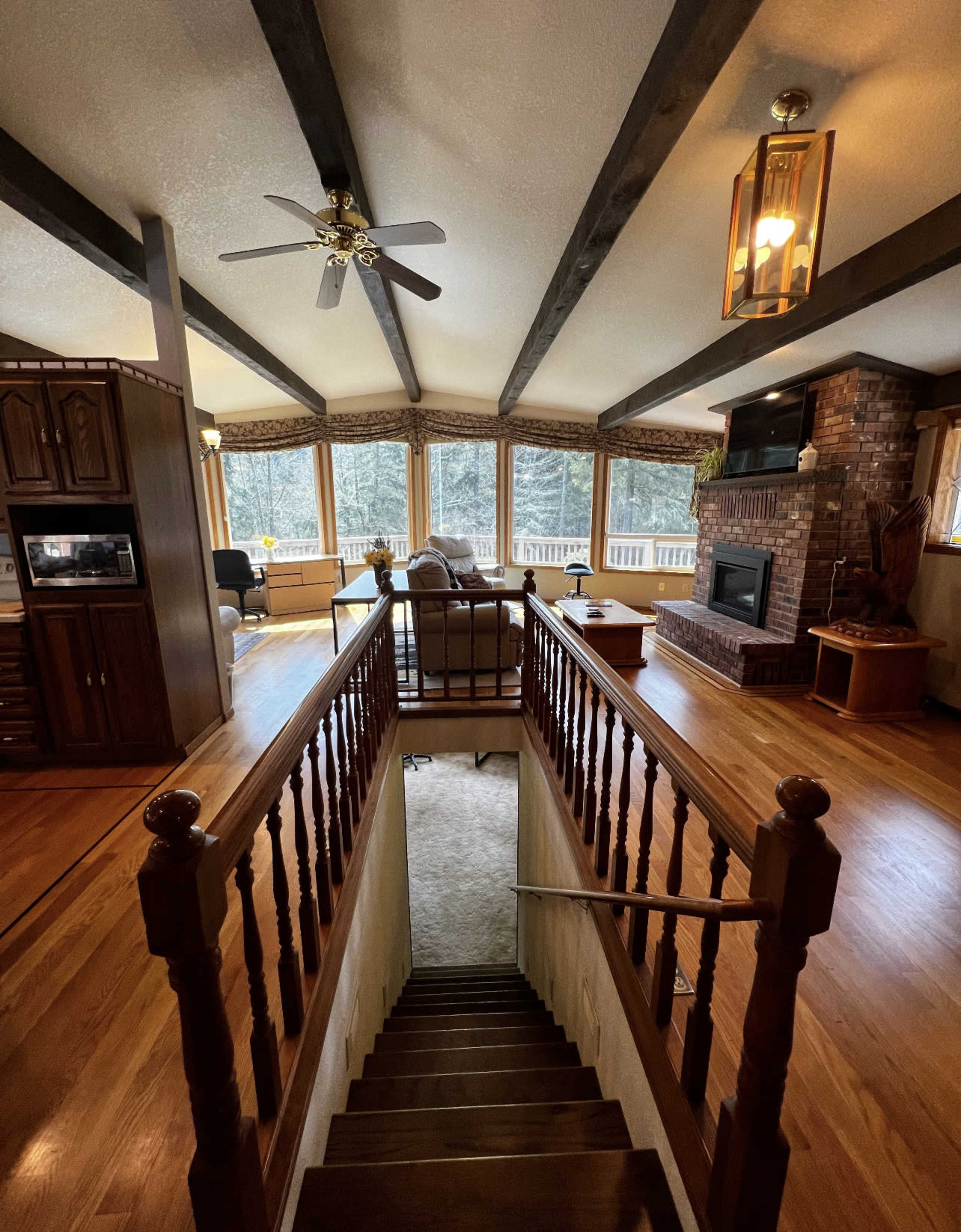 A wooden staircase leads down to a spacious living area with large windows and exposed beams, featuring a fireplace and seating arrangement.
