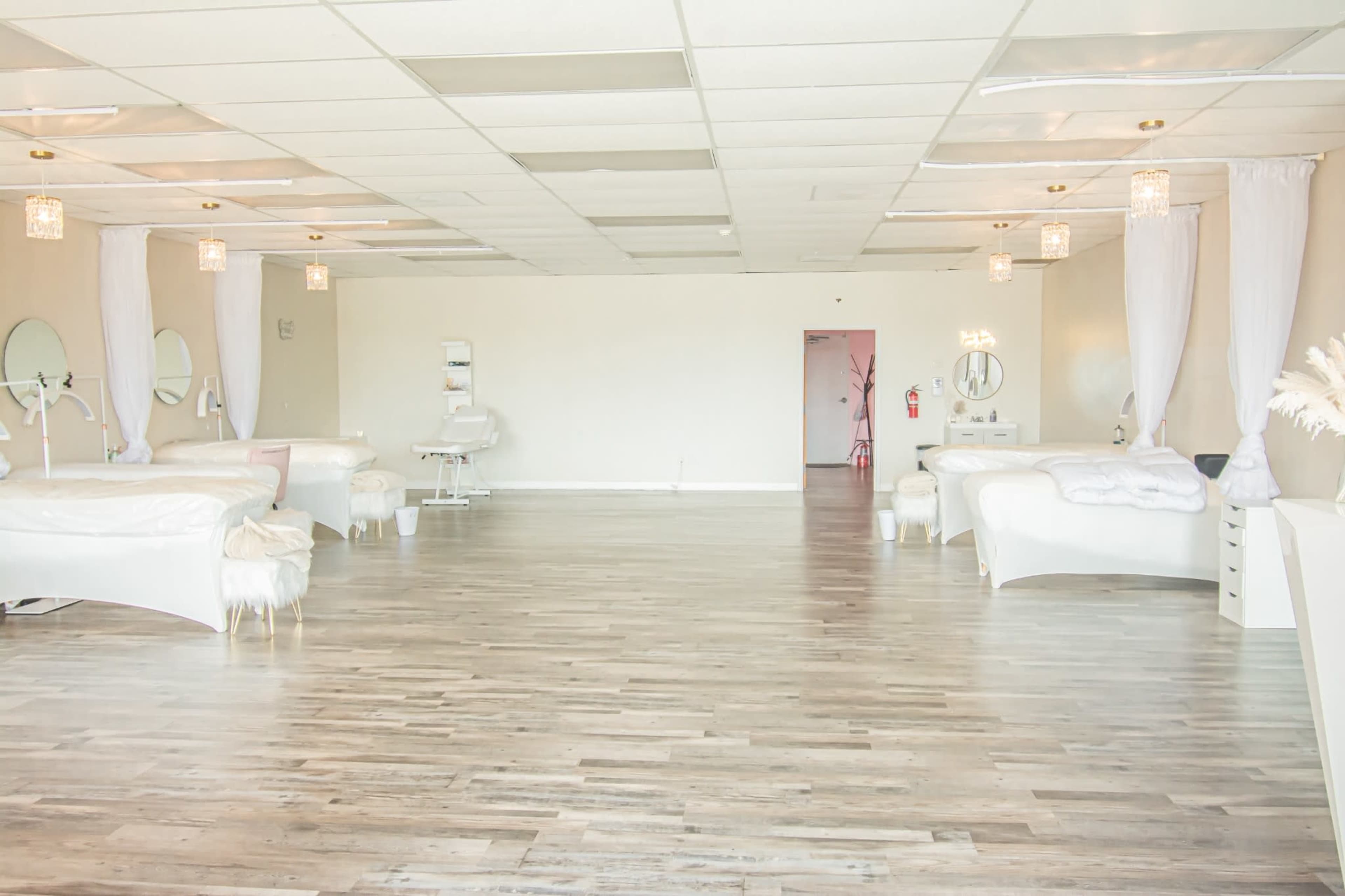 The image shows a spacious and well-lit room with white décor, featuring multiple beauty treatment stations and large mirrors.