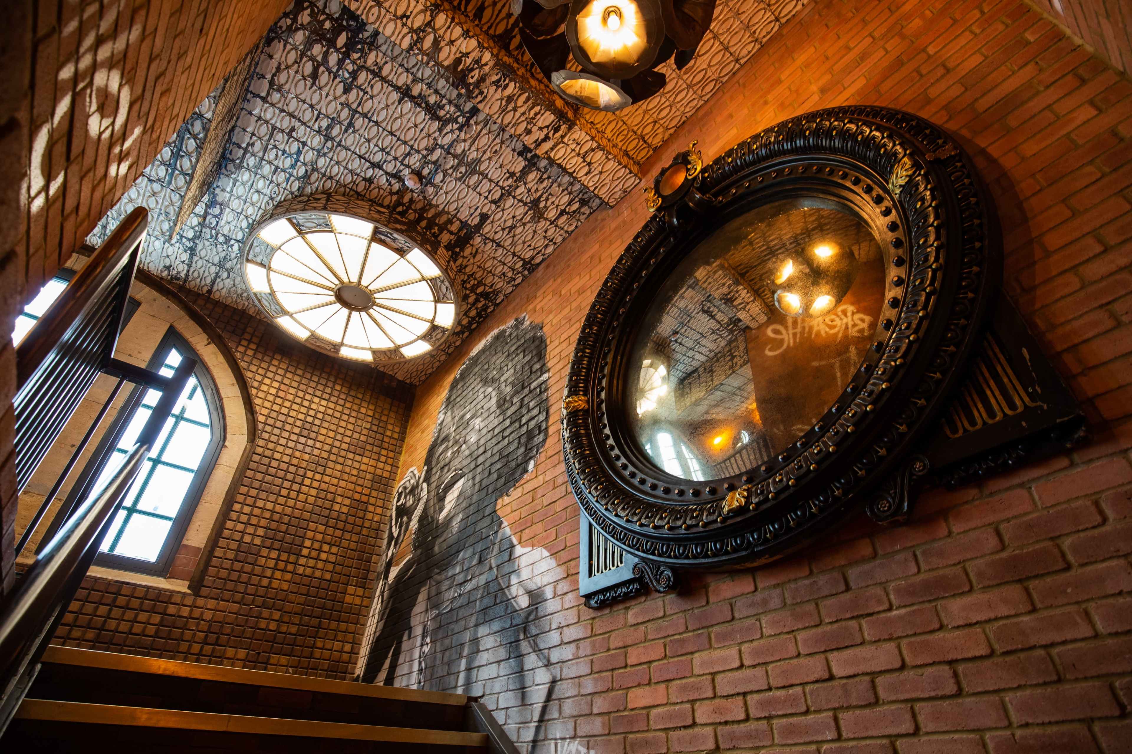 The image shows a staircase with brick walls, a large decorative mirror, and a round window above, featuring a painted mural on the ceiling.