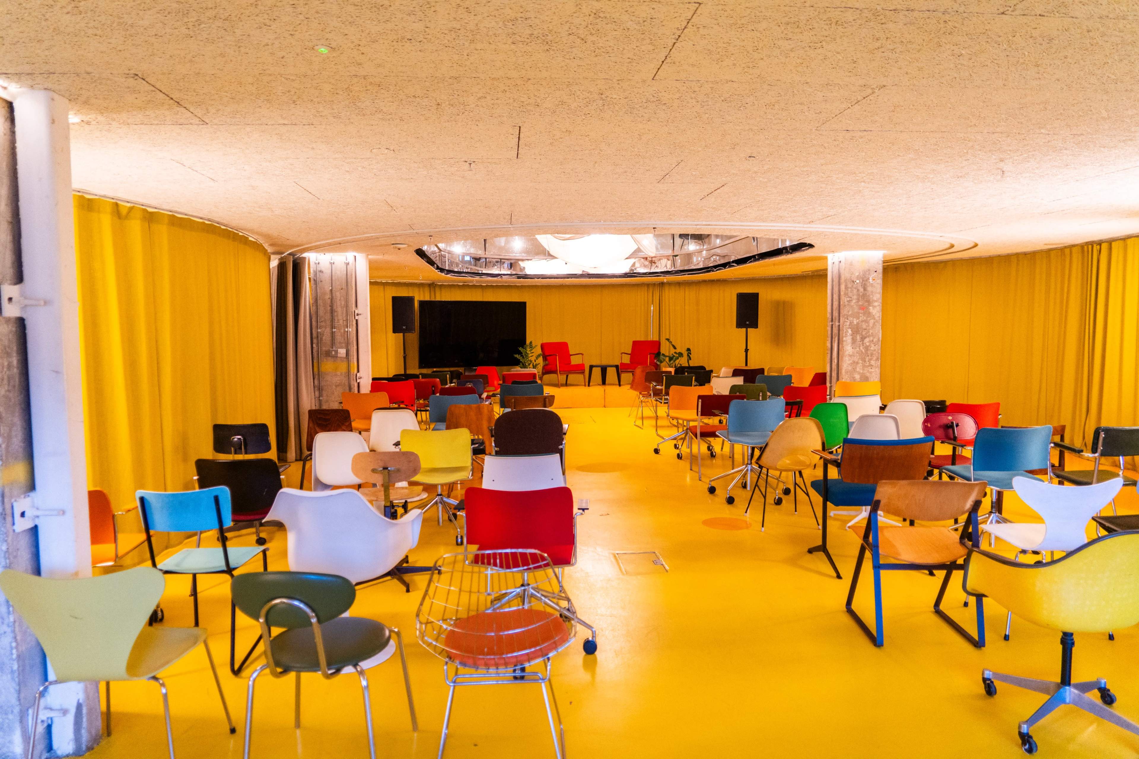 The image shows an empty circular room with colorful chairs arranged in rows on a bright yellow floor under a ceiling with indirect lighting.