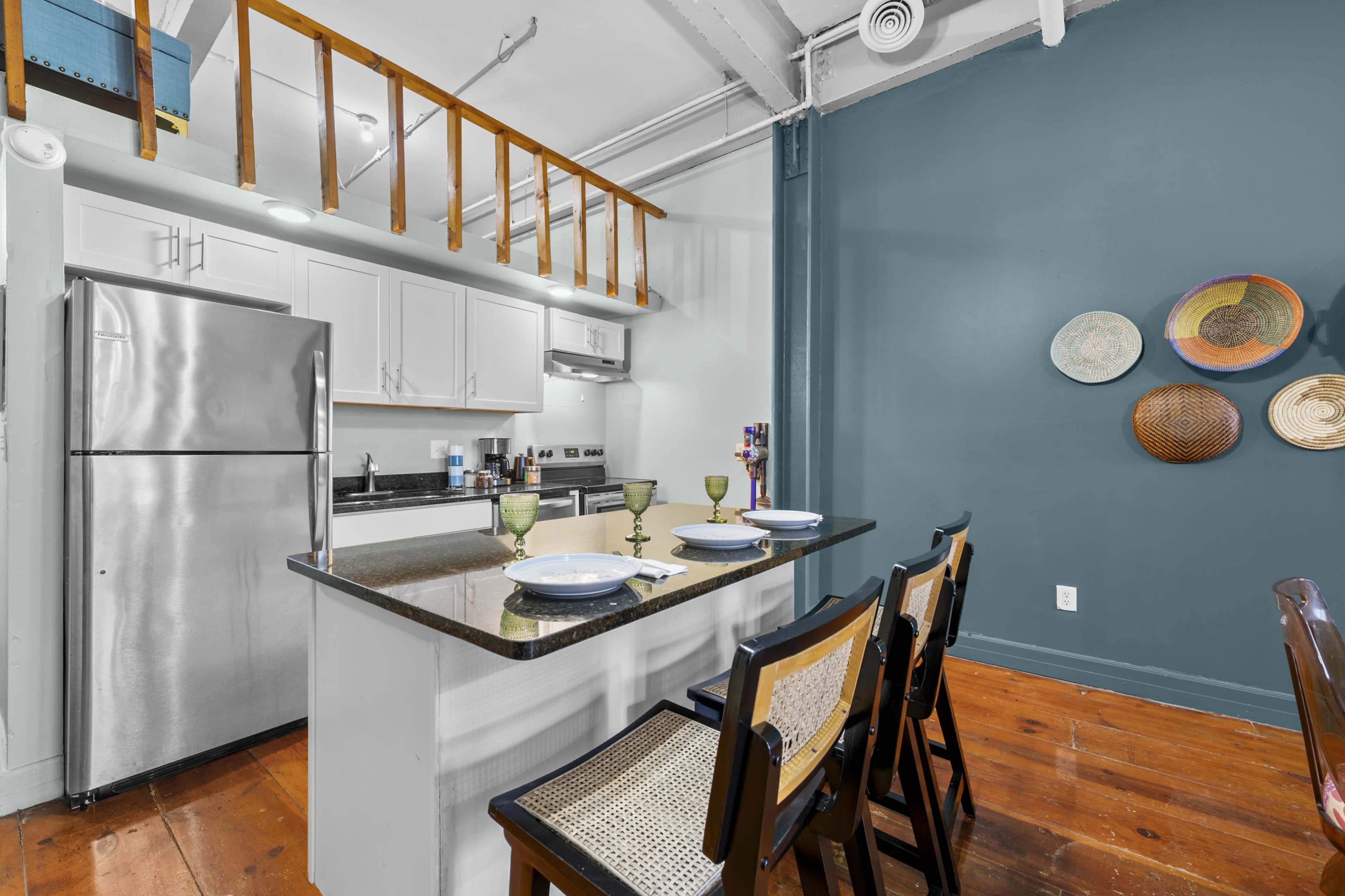 The image shows a modern kitchen with white cabinets, a stainless steel refrigerator, and a dining area with bar stools and decorative plates on the wall.