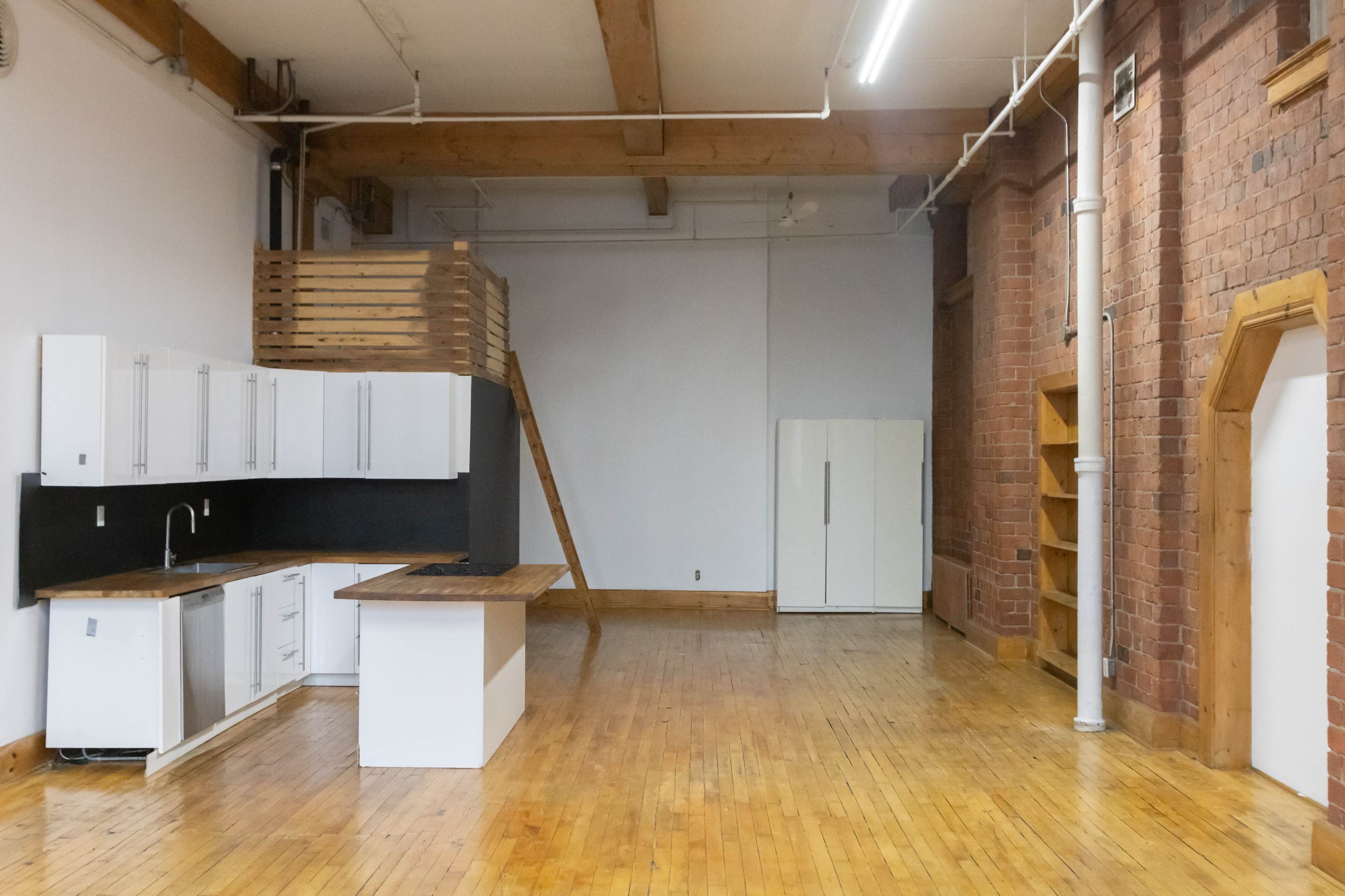 The image shows an open loft space featuring a kitchen area with white cabinets, a wooden countertop, and a refrigerator, along with exposed brick walls and wooden beams.