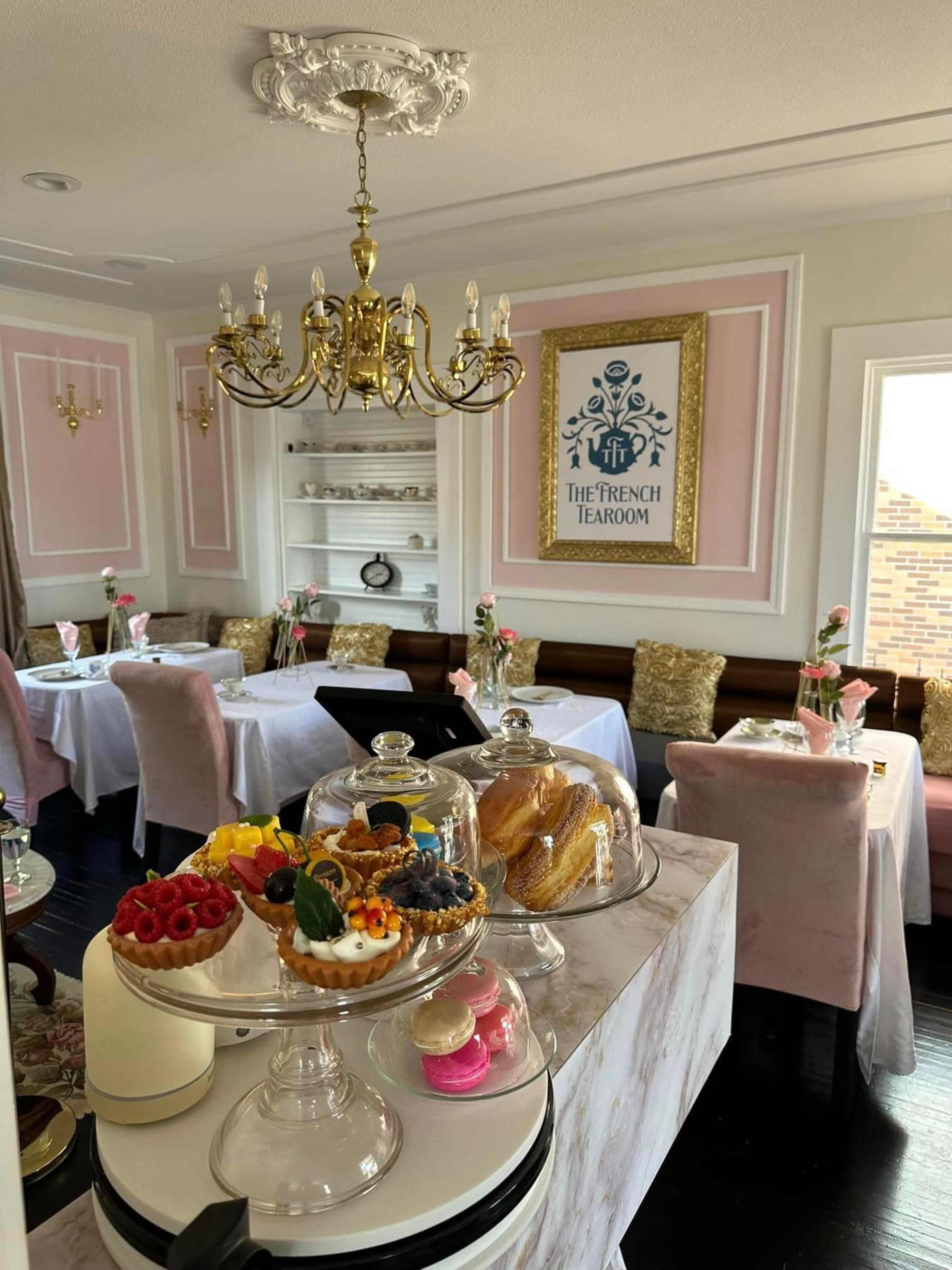 A beautifully arranged tearoom features elegant tables with pink and white decor, surrounded by a chandelier and showcasing an array of desserts on display.