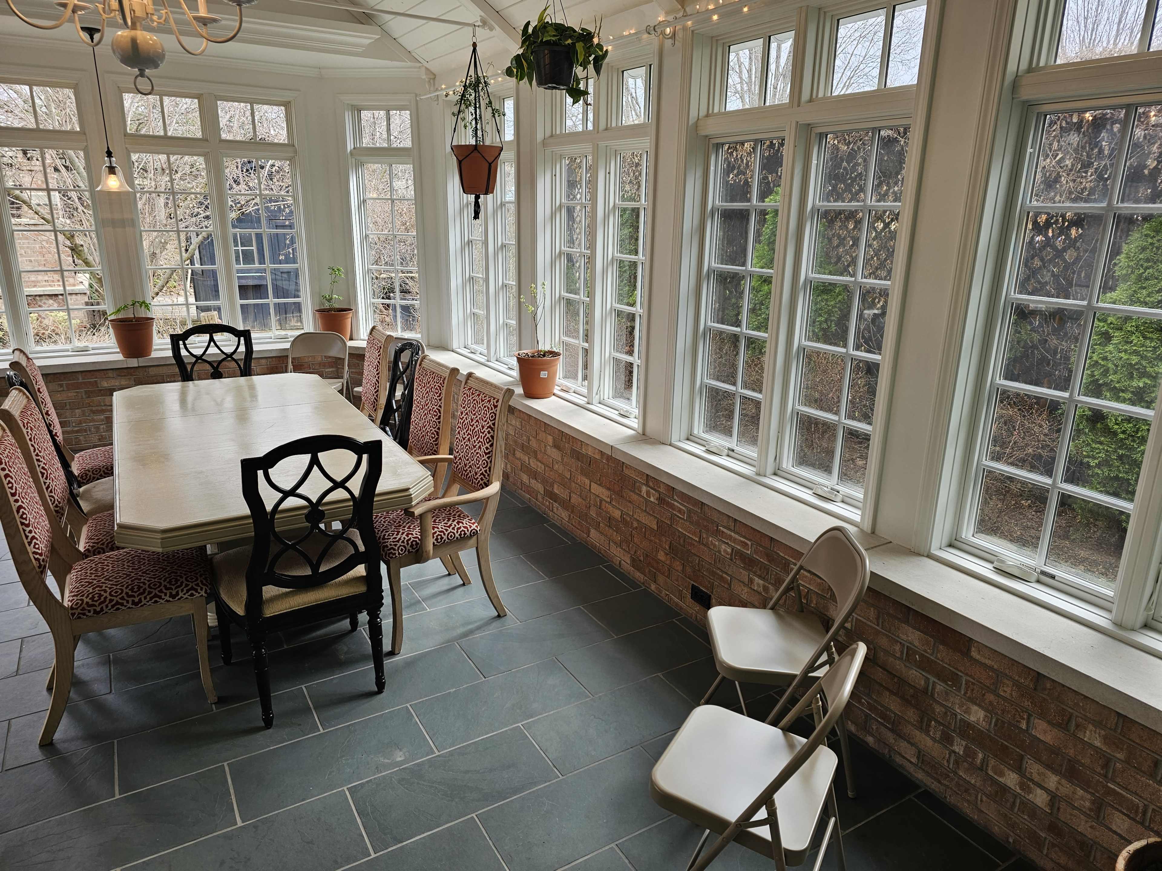 A sunlit dining room features a large table surrounded by chairs, with large windows showcasing a garden view.