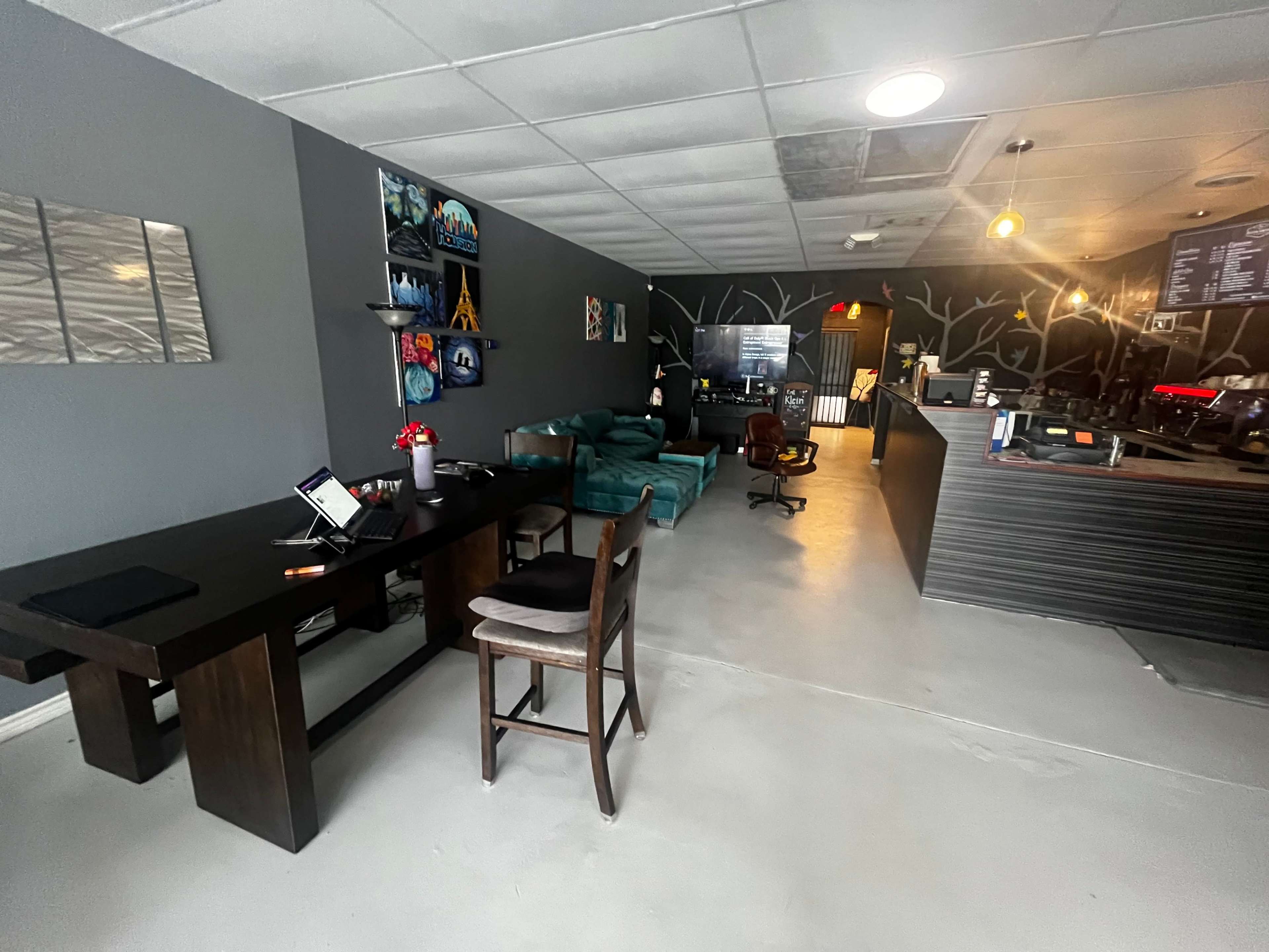 The image shows a modern, casual interior of a café with furniture including a dark wood table, chairs, and a teal couch against gray walls adorned with artwork.