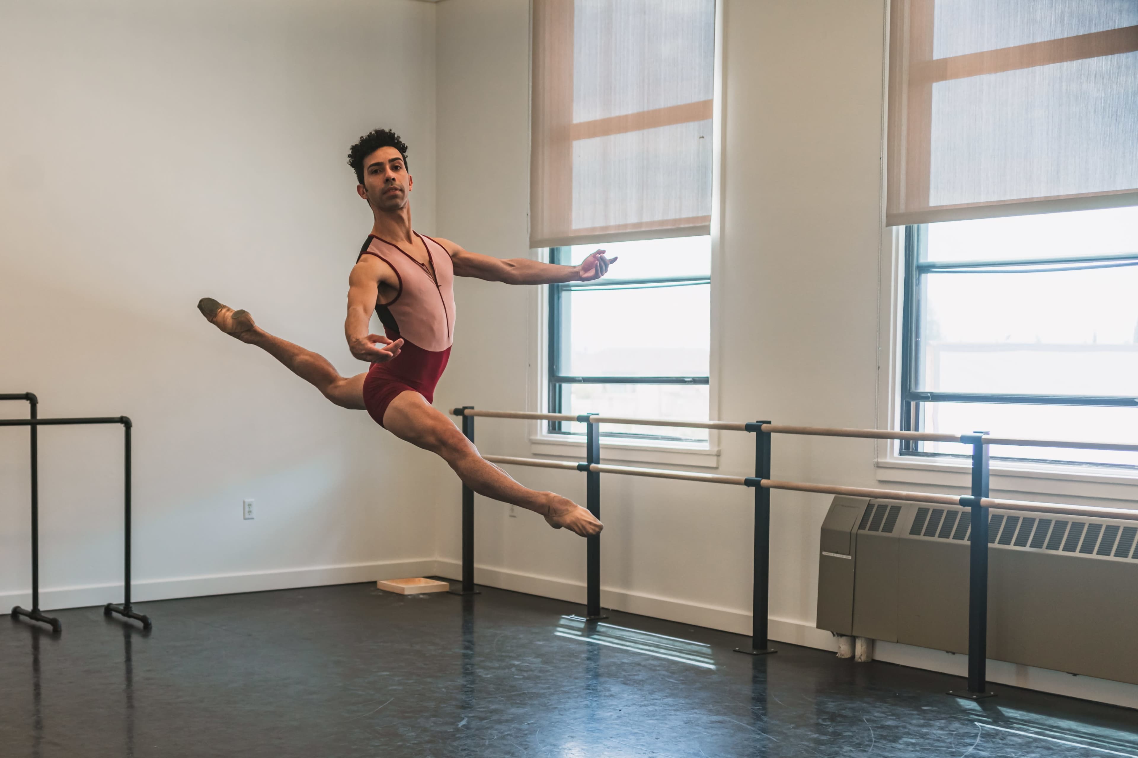A male ballet dancer performs a leap in a spacious dance studio with large windows and a wooden floor.