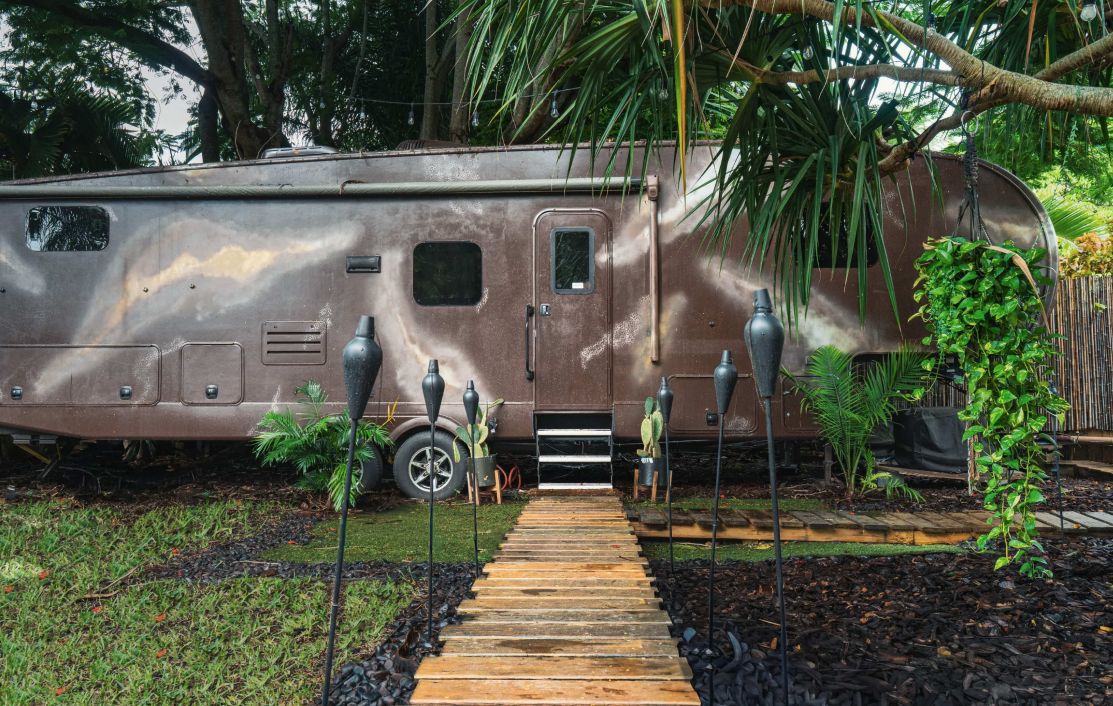 A weathered trailer with a narrow wooden walkway leads to its entrance, surrounded by tropical plants and landscaping.