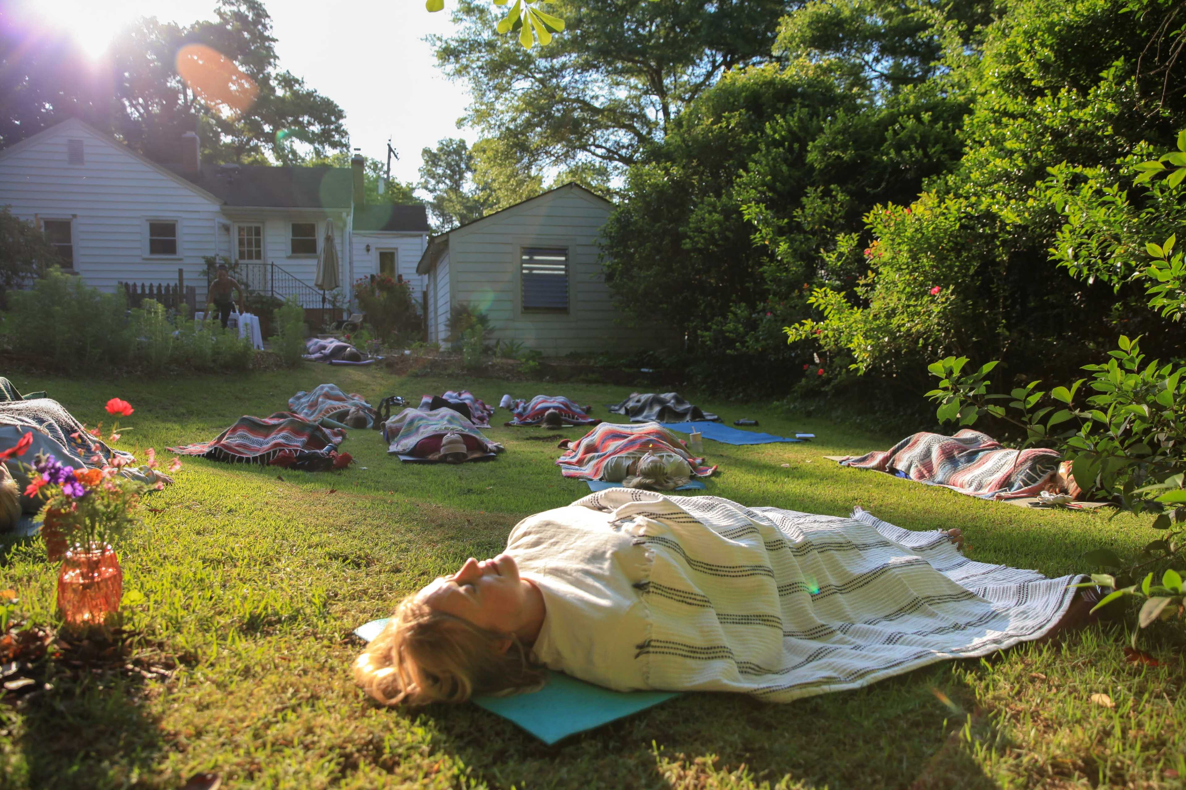 A group of people lies on mats in a grassy area, wrapped in blankets, with a house and trees in the background.