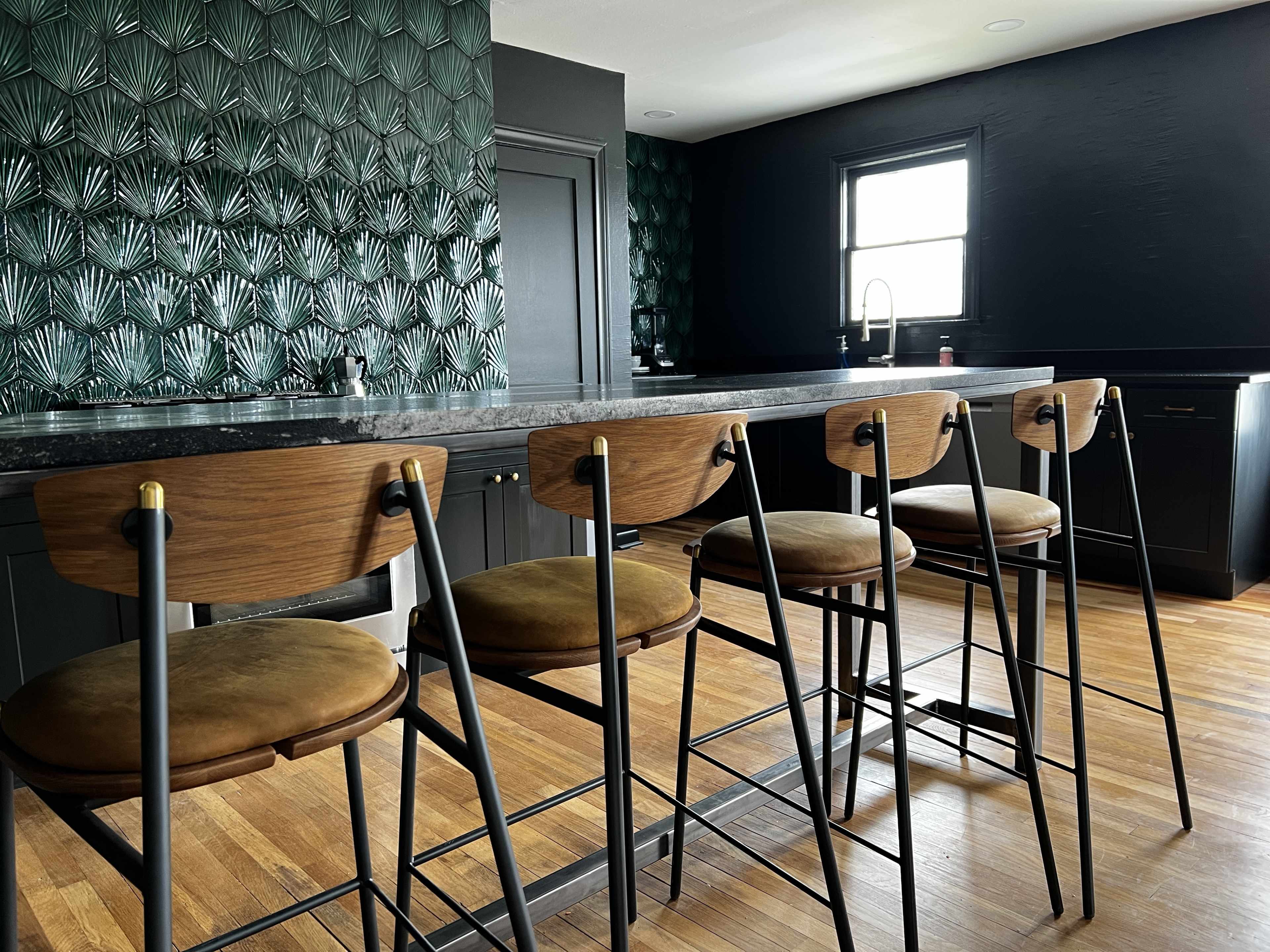A sleek kitchen features a black wall with textured green wallpaper, a long bar with four wooden stools, and modern cabinetry.