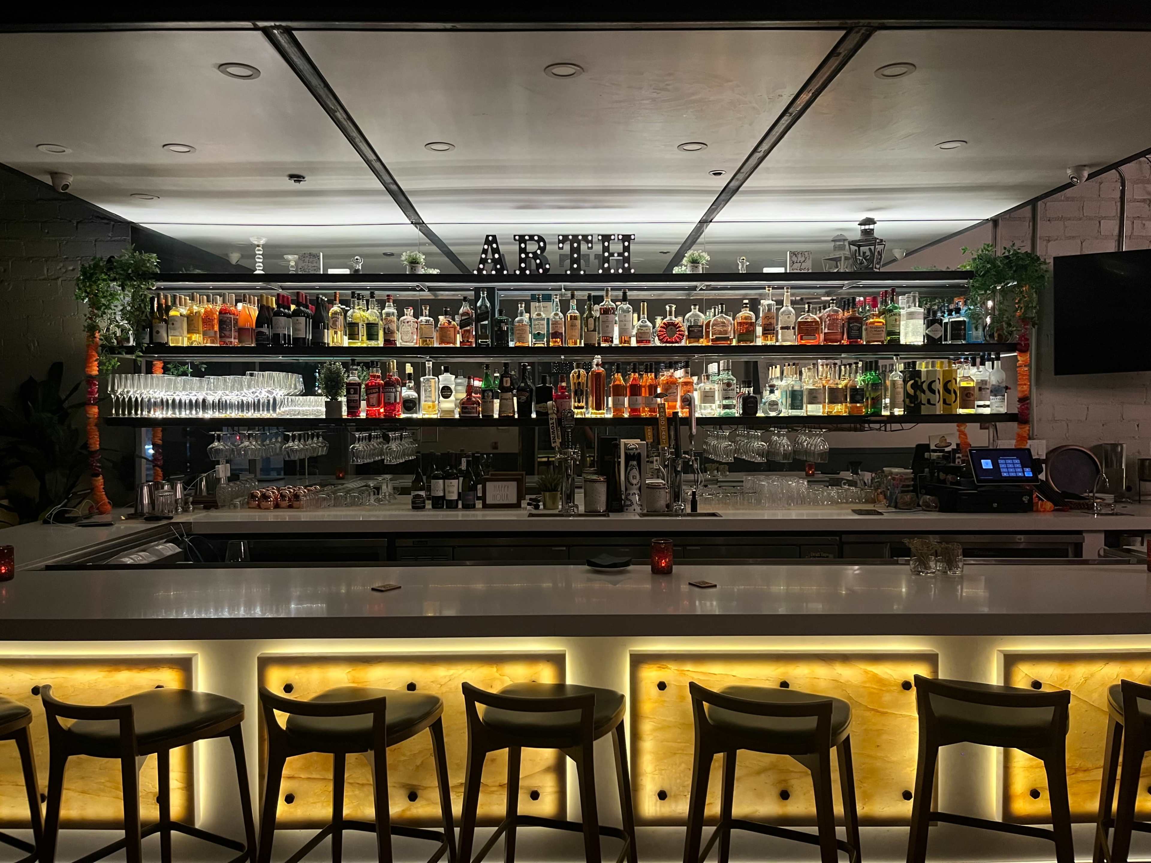A well-lit bar features a variety of liquor bottles displayed on shelves behind a sleek countertop with seating in front.