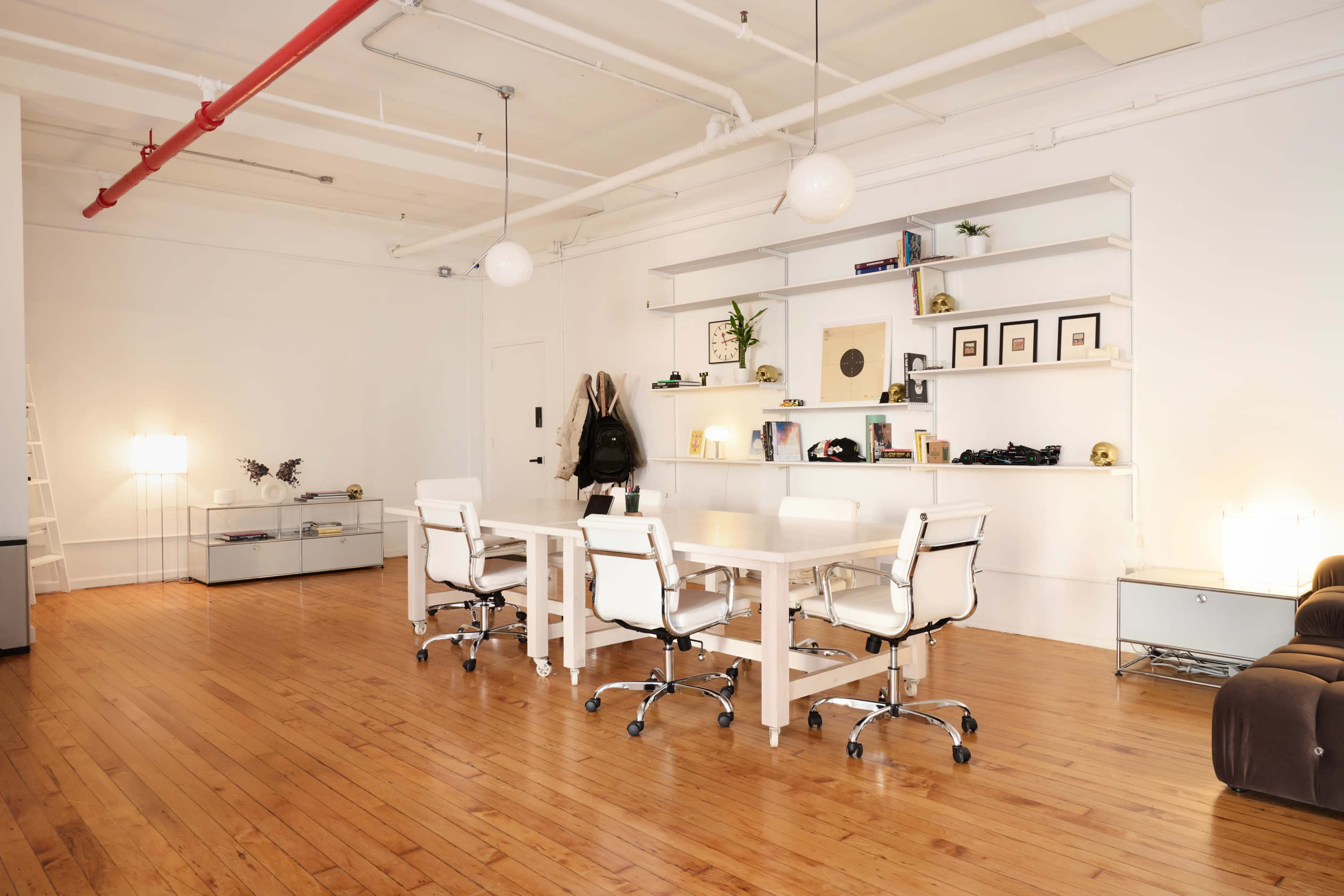 A bright, open office space features a large white table surrounded by ergonomic chairs, with shelves displaying framed photos and decorative items along the wall.