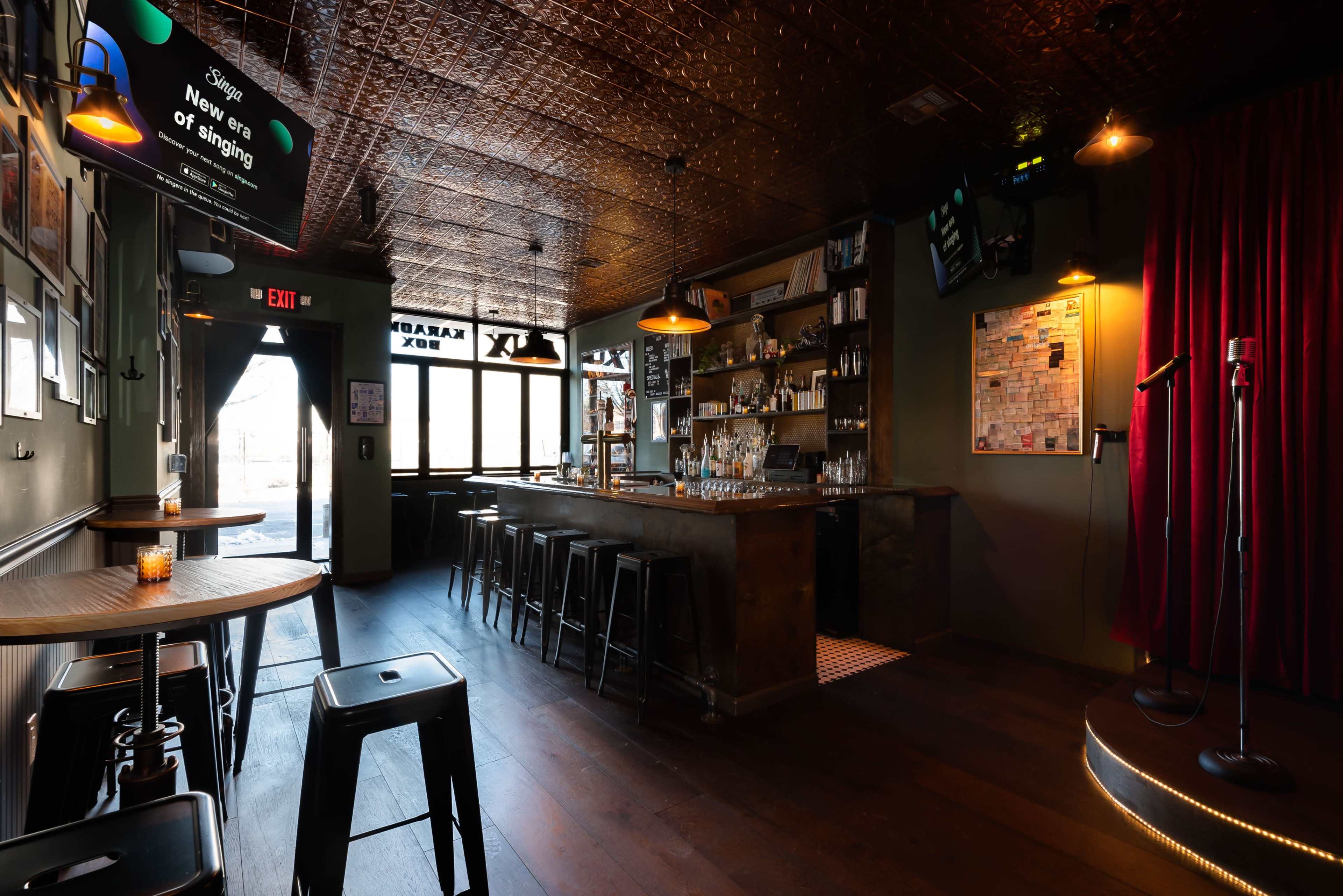 The image shows a dimly lit bar with a wooden floor, a long counter with stools, and a small stage with a microphone.
