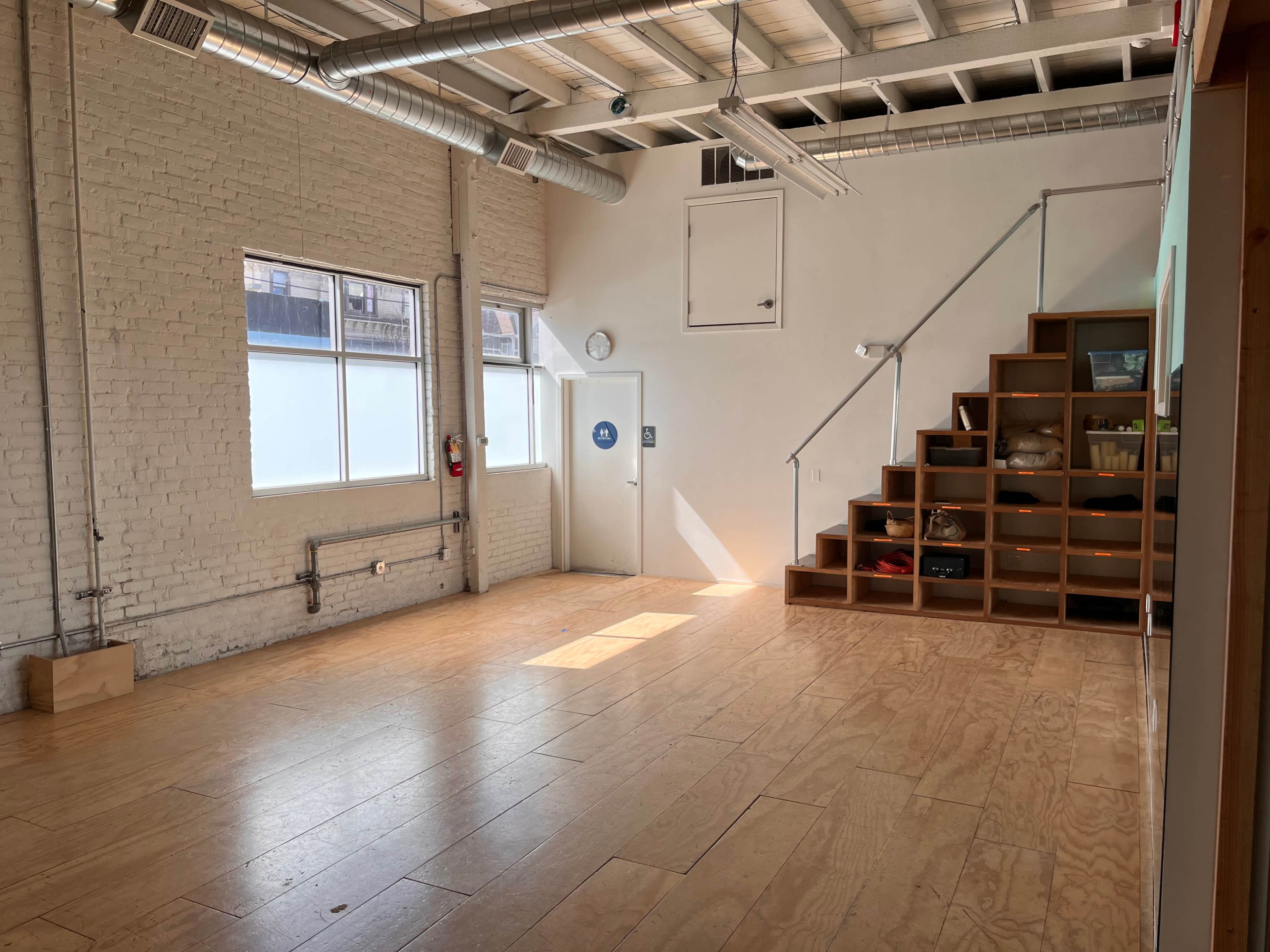 A spacious interior room with wooden flooring, large windows, and a staircase leading to a storage area along one wall.