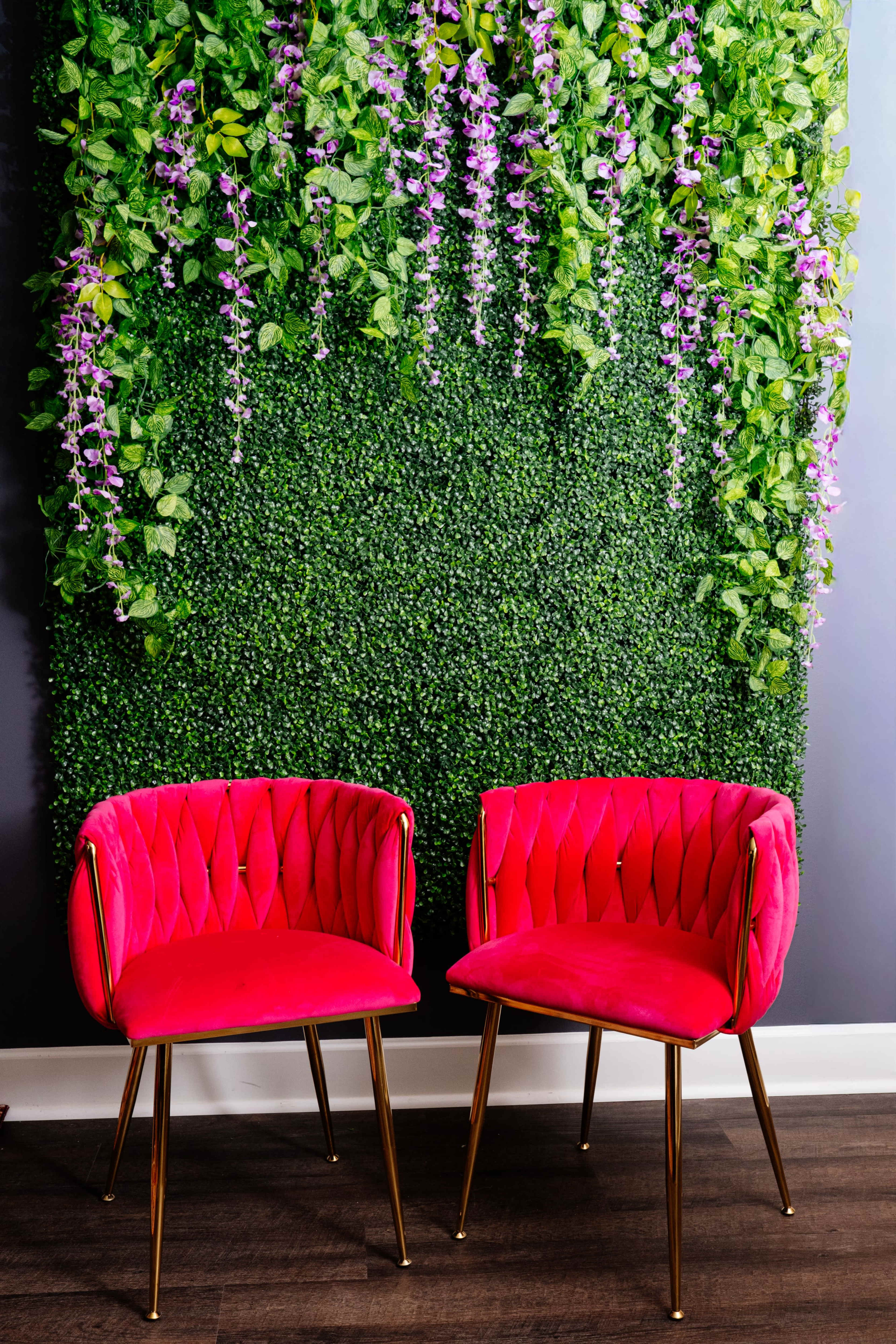 Two pink velvet chairs with gold legs are positioned in front of a vertical garden wall adorned with green foliage and purple flowers.
