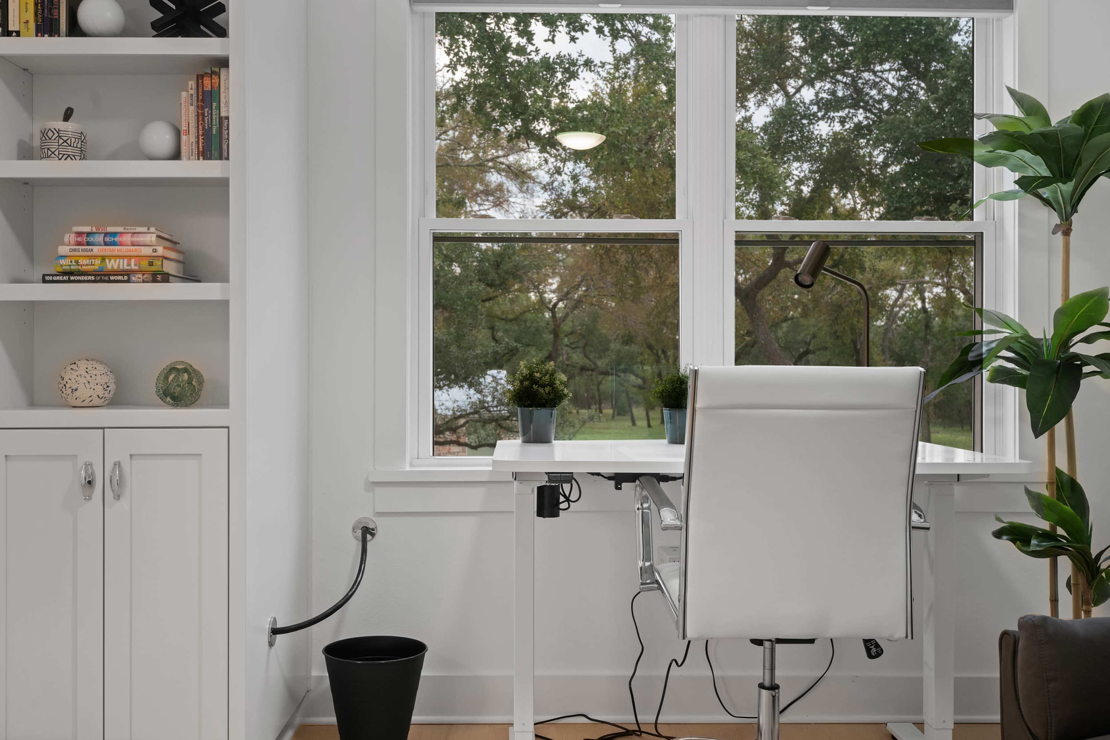 A white desk and chair are positioned near a window with a view of trees outside, complemented by a bookshelf filled with books and decorative items.
