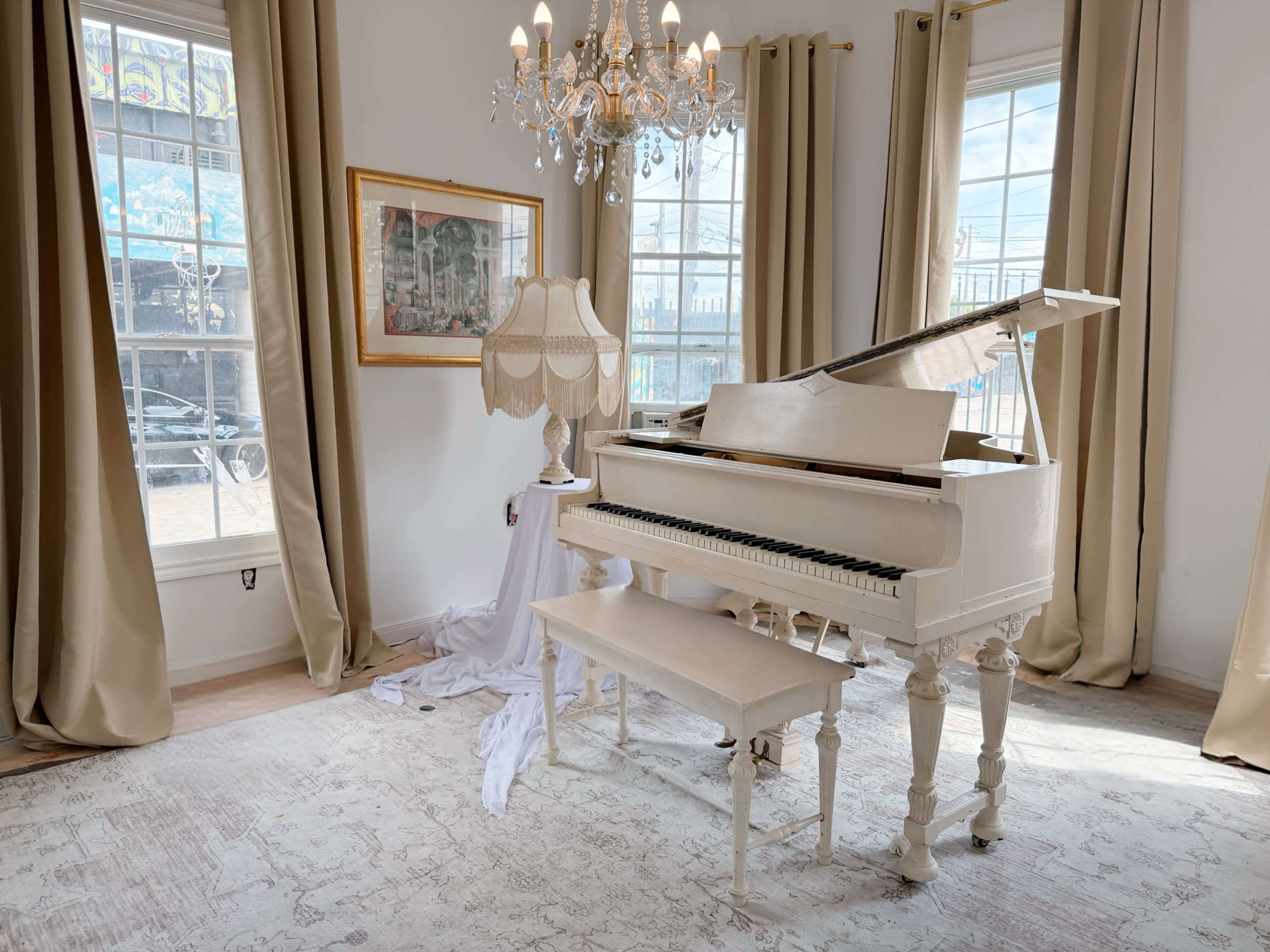 A white grand piano is positioned in a bright room with large windows, a chandelier, and draped curtains.