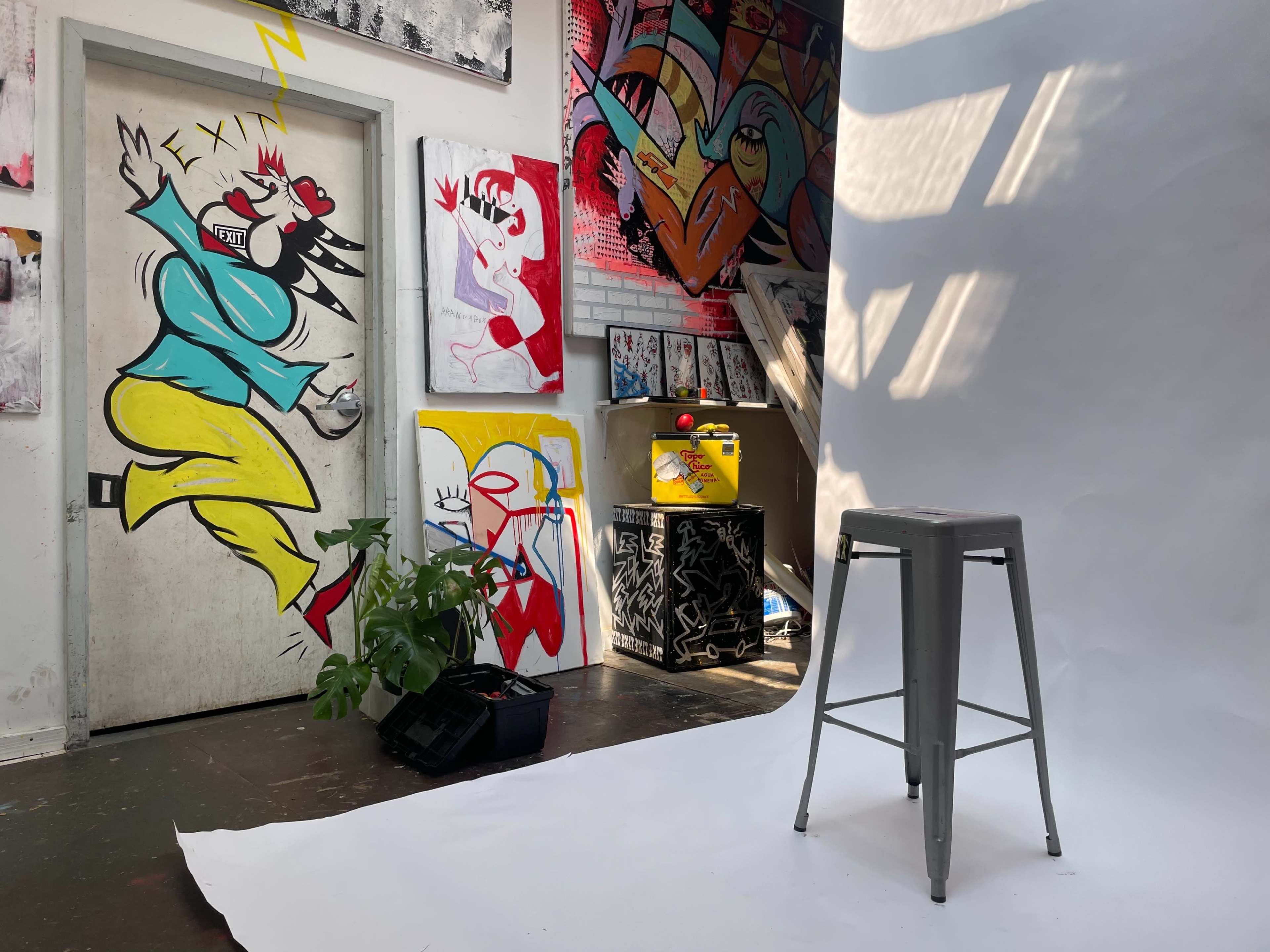 An art studio features a vibrant mural on the wall, a bare white backdrop in the foreground, and a metal stool beside potted plants and a decorated cabinet.