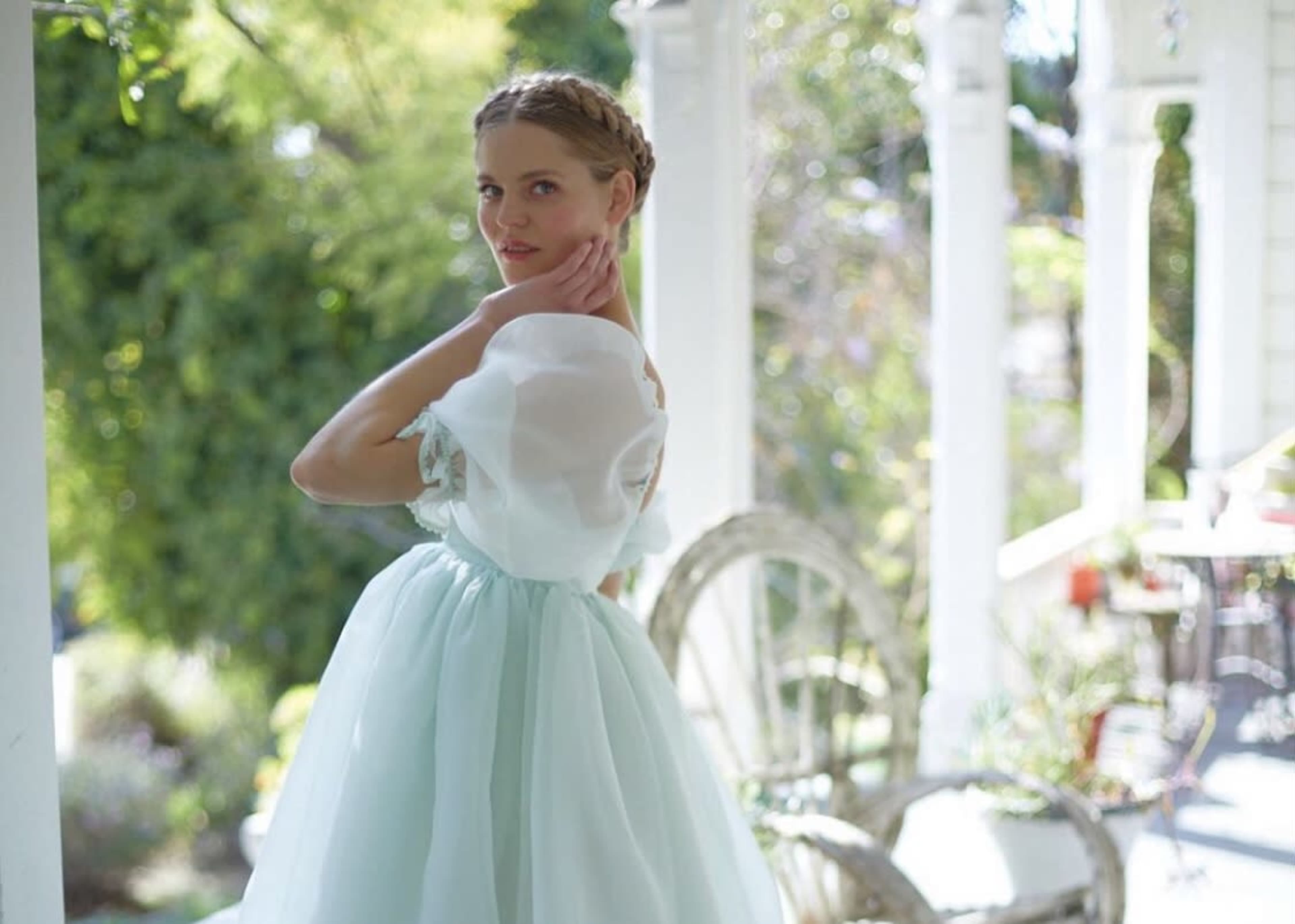A woman in a light blue dress poses outdoors near a porch surrounded by greenery.