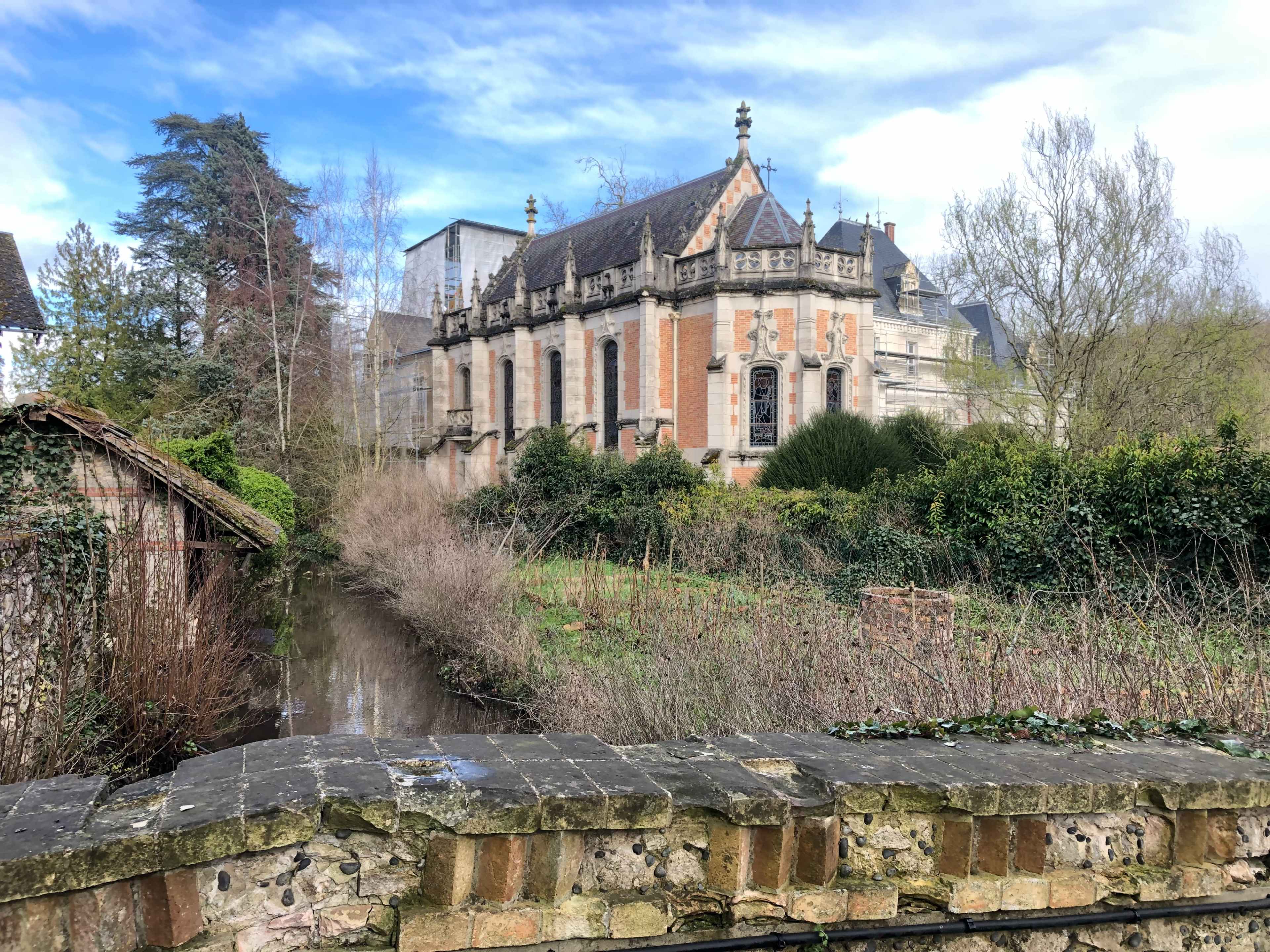 A brick building with a steep roof stands next to a serene, overgrown waterway, surrounded by bare trees and shrubs.