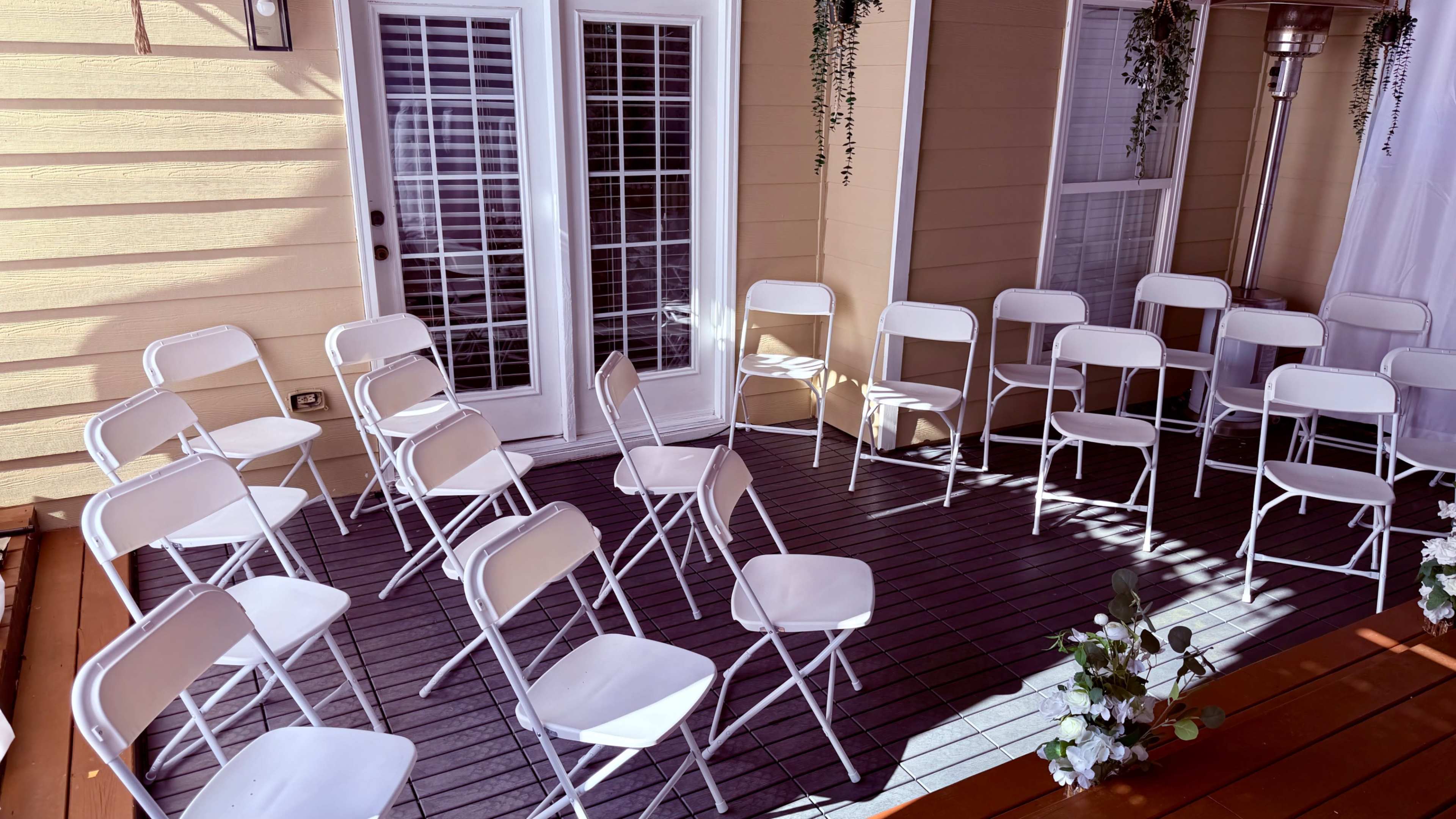 The scene shows a set of white folding chairs arranged in a circular formation on a deck next to large glass doors.