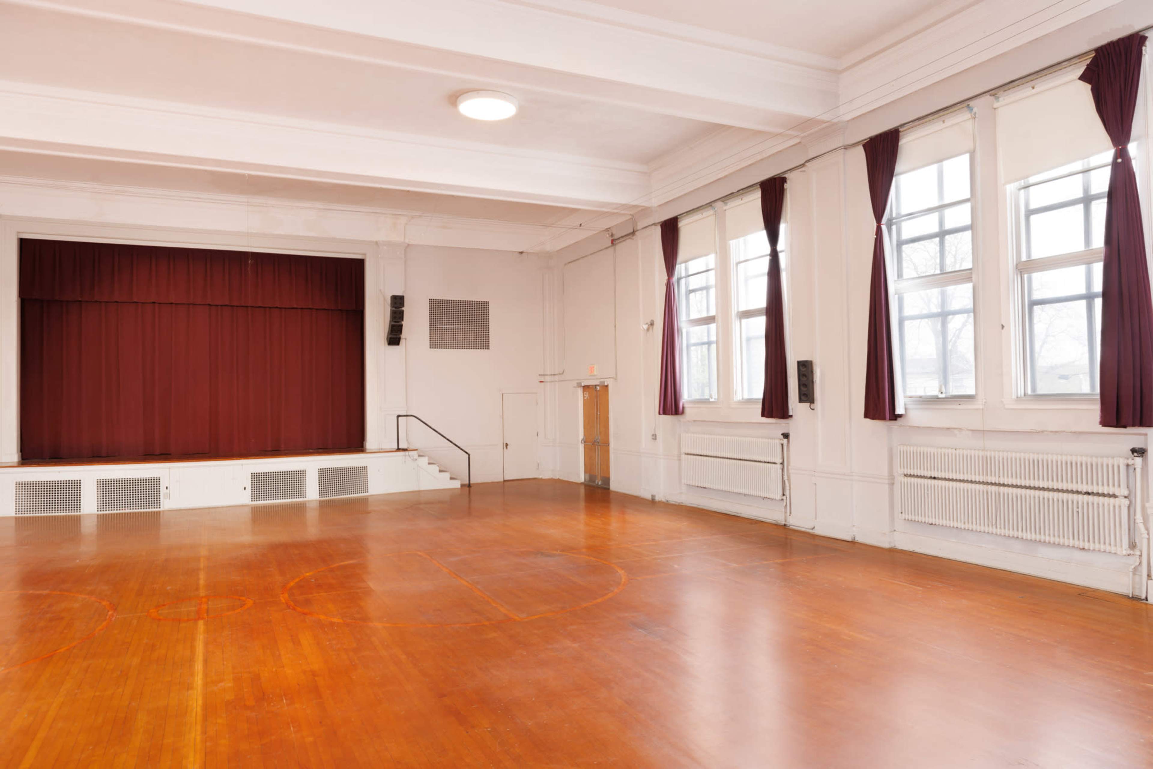 An empty gymnasium with a polished wooden floor, large windows, and a stage draped in red curtains.