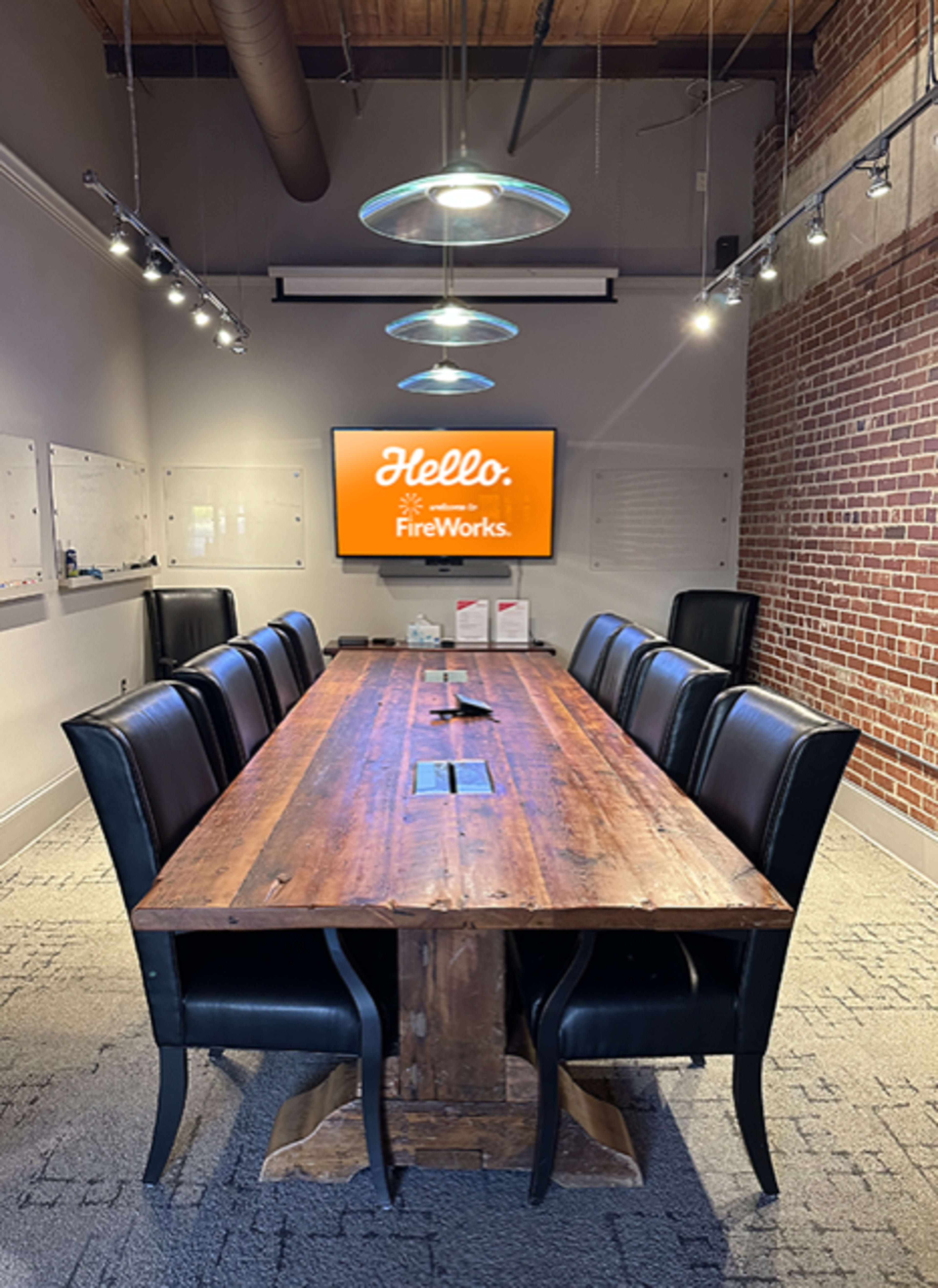 A large wooden conference table with black leather chairs is centered in a meeting room featuring a wall-mounted screen displaying "Hello, FireWorks."