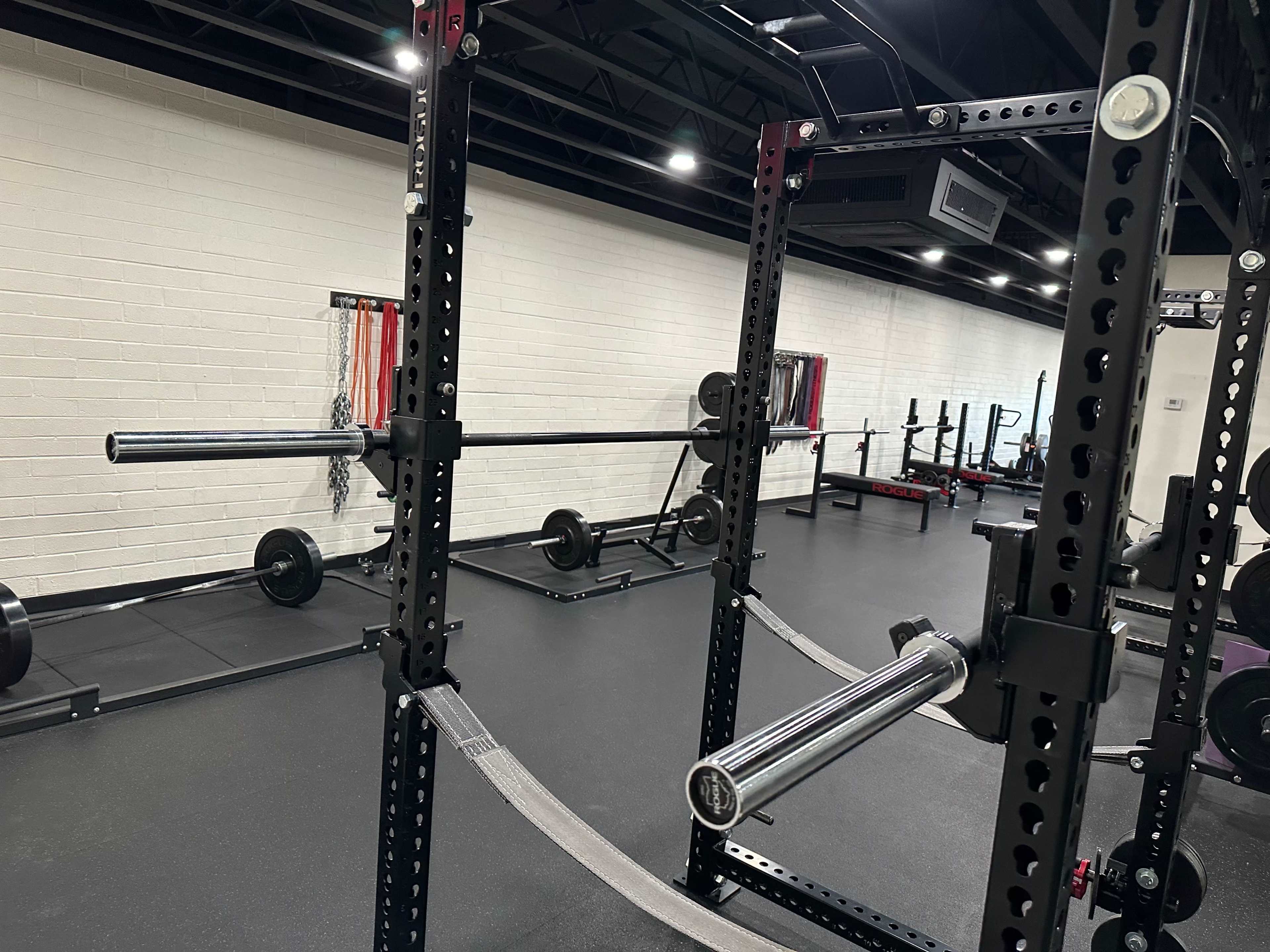 The image shows a well-equipped gym space featuring a squat rack, barbells, and weight plates on the floor.