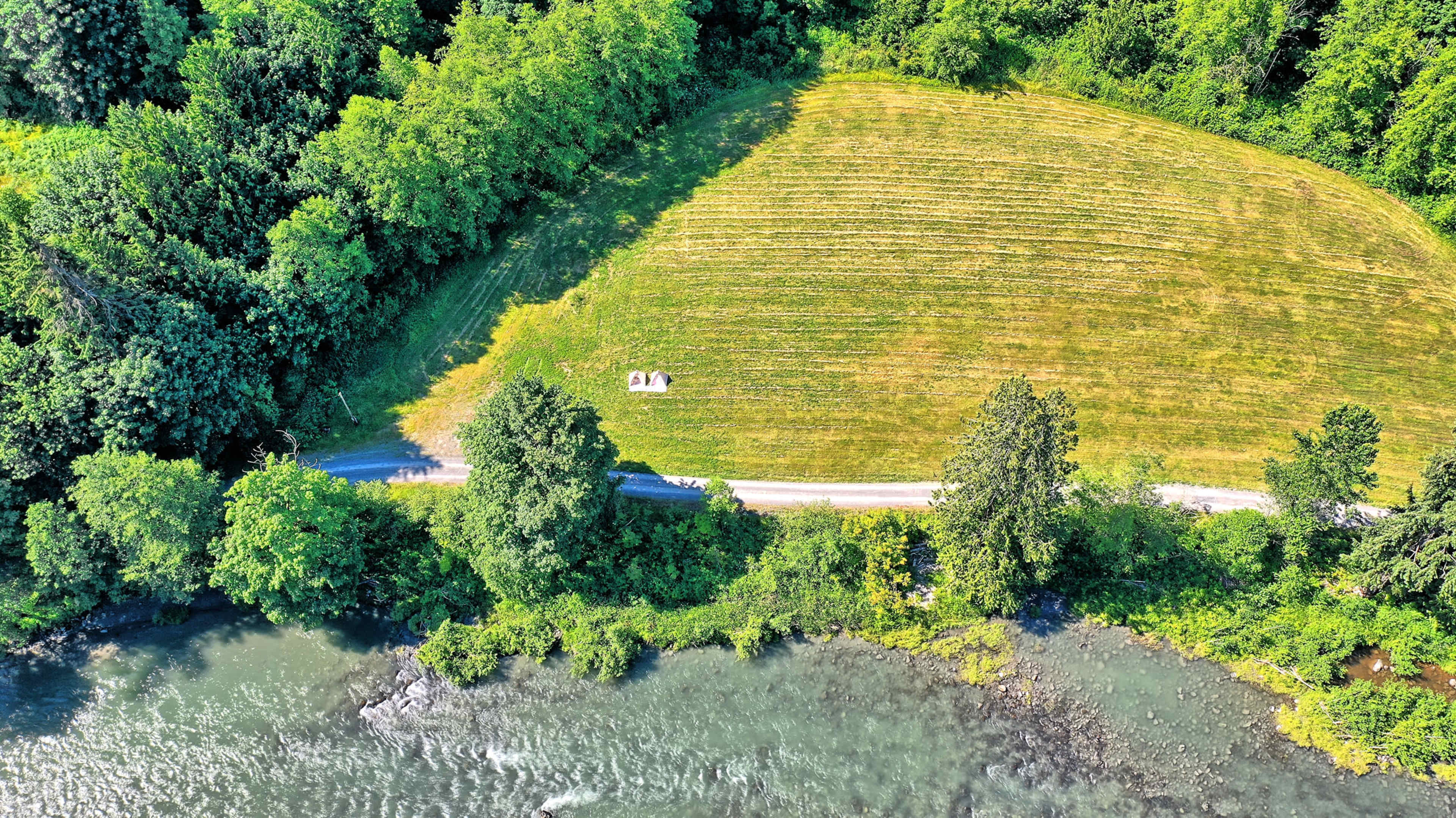 A green landscape with a river at the bottom and a neatly mowed field bordered by trees, with a small structure near the edge of the field along a dirt road.