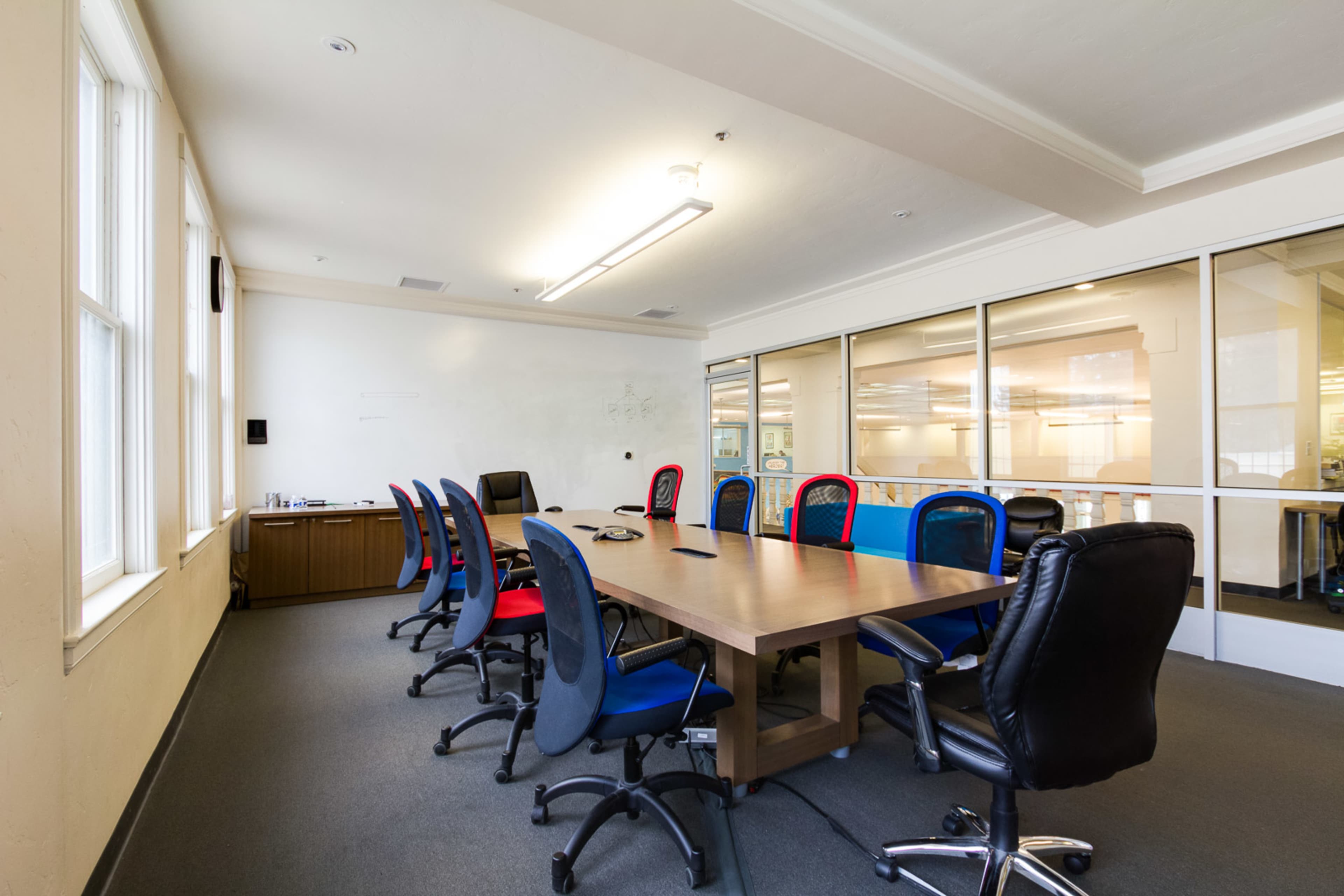 A conference room features a large wooden table surrounded by chairs in red and blue, with windows providing natural light.