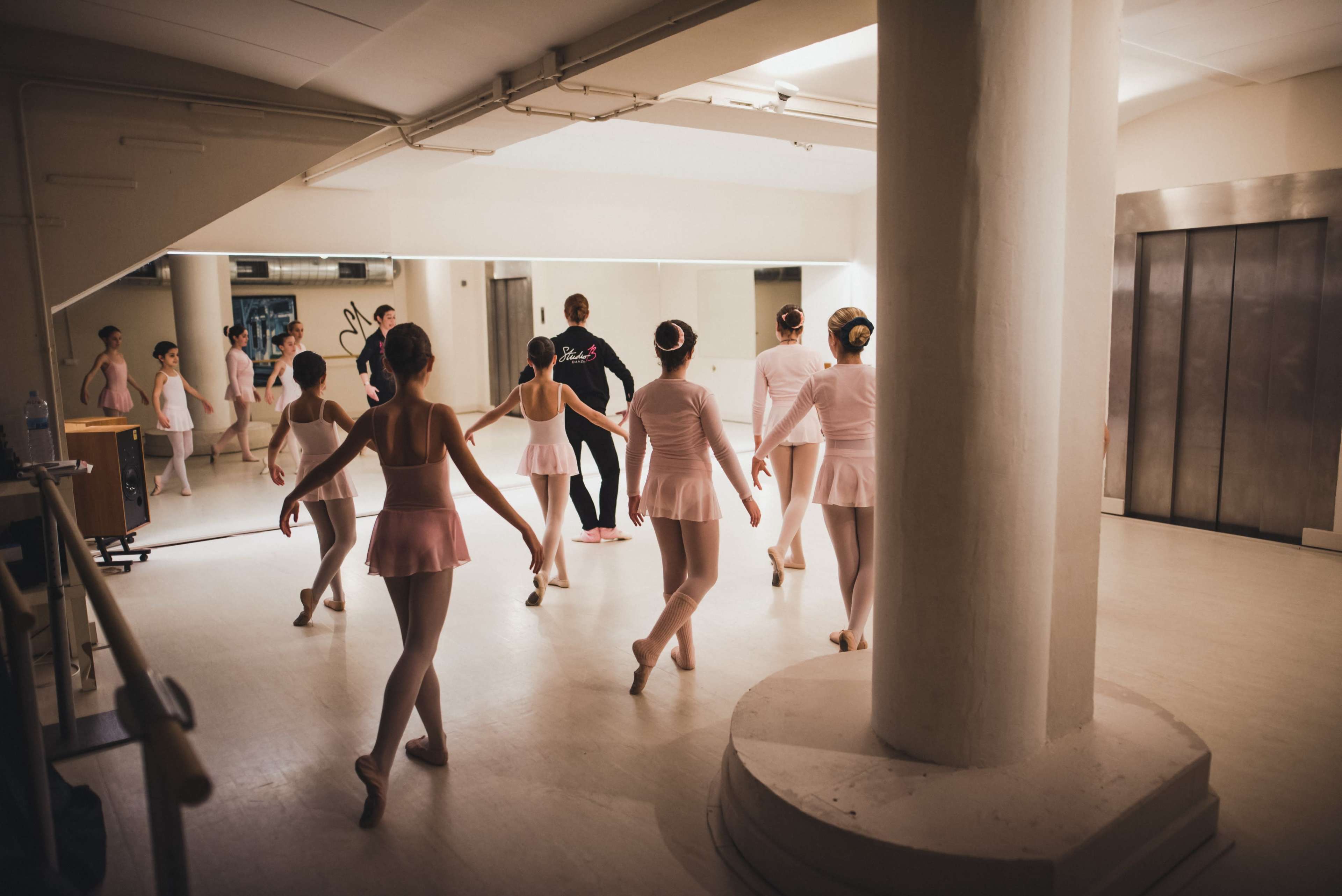 A group of ballet dancers in pink costumes practice in a mirrored studio adjacent to an elevator.