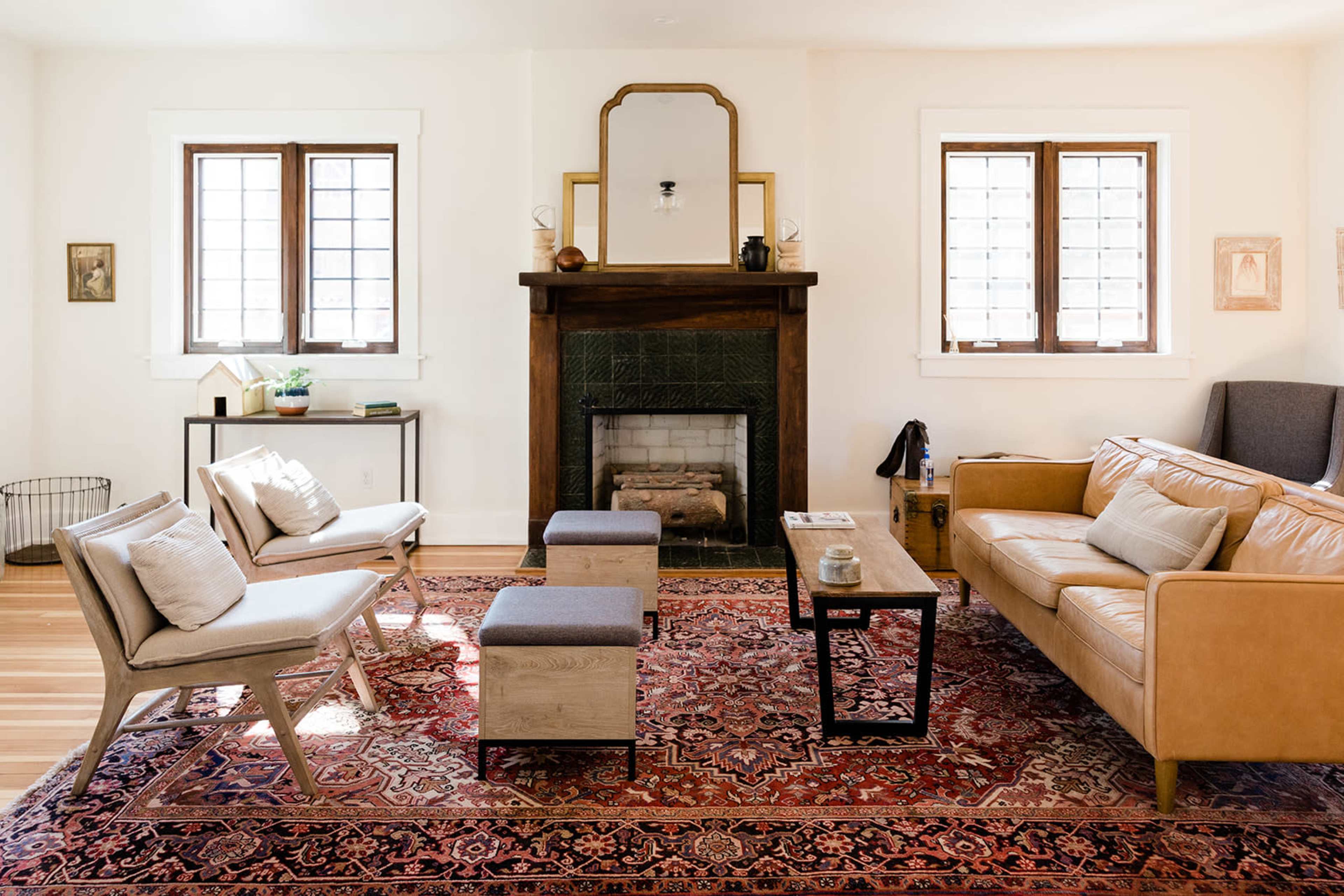 The room features a cozy living area with a leather sofa, two armchairs, and a coffee table arranged around a patterned rug, with a fireplace and windows providing natural light.