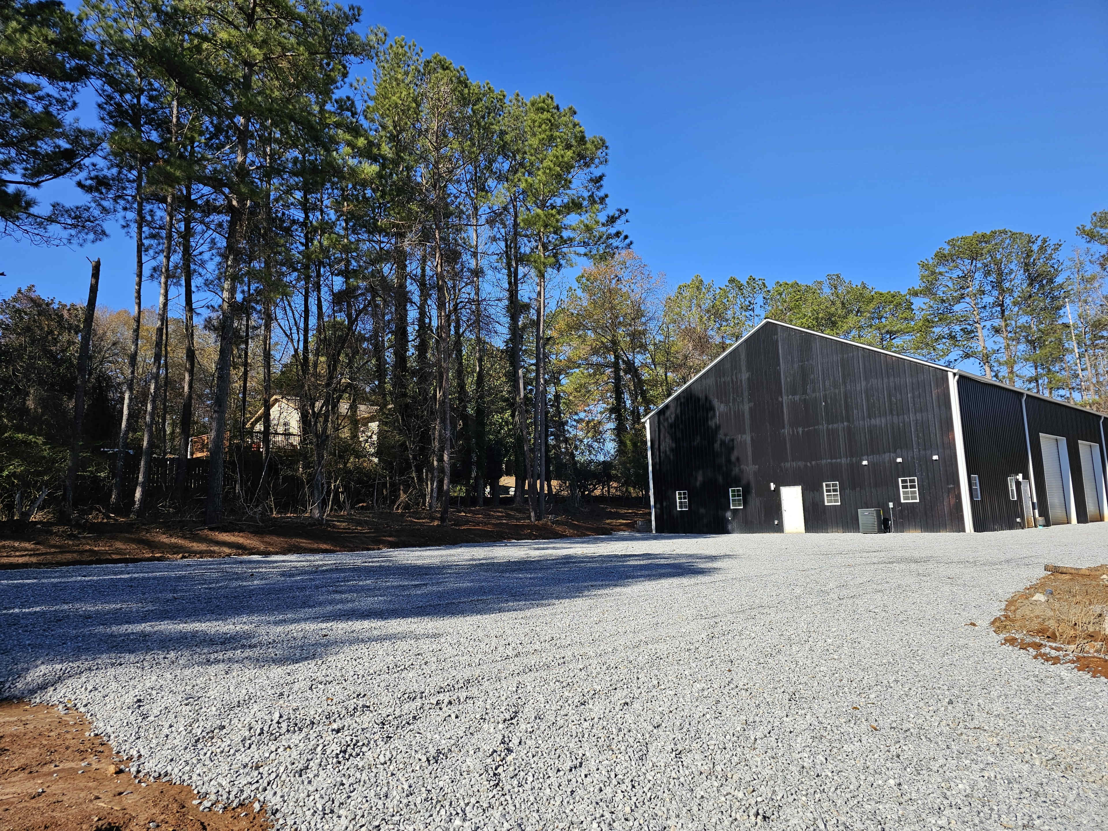 A gravel driveway leads to a large black barn surrounded by tall pine trees under a clear blue sky.