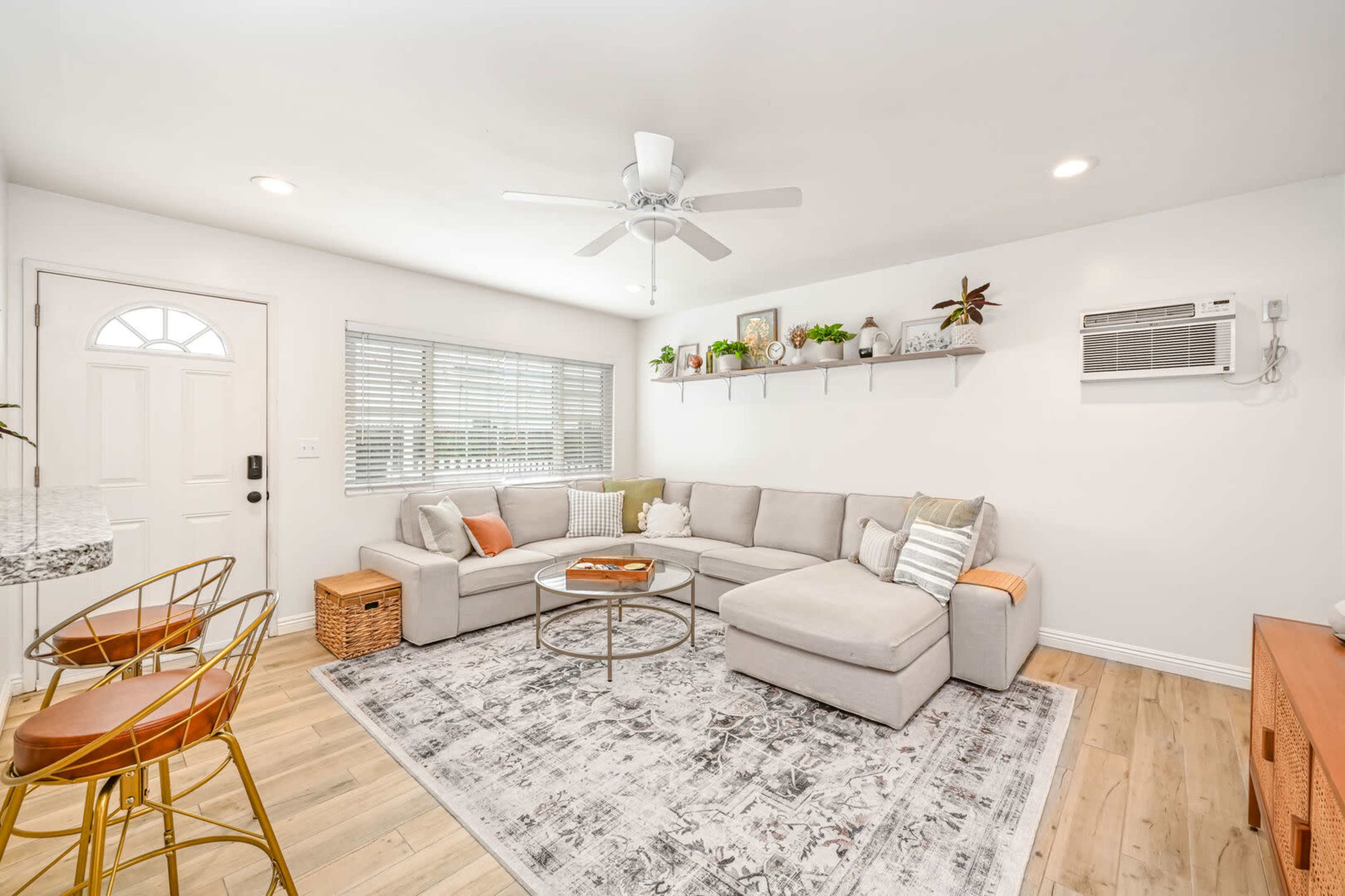 A light and airy living room features a gray sectional sofa, a round coffee table, and a wall-mounted shelf with plants, alongside a front door and a window with blinds.