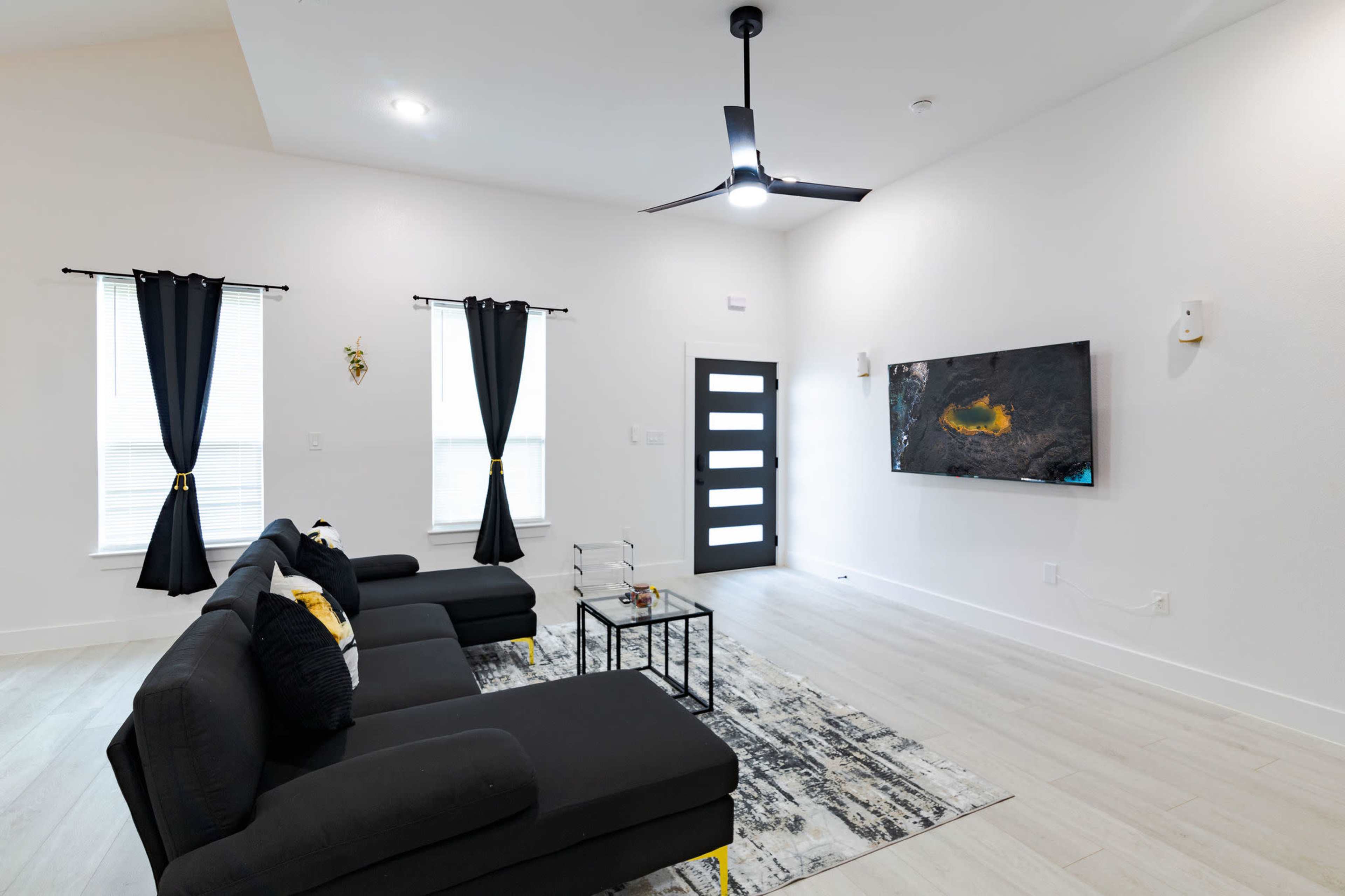 The living room features a black sectional sofa, a modern ceiling fan, a wall-mounted television, and large windows with black curtains.