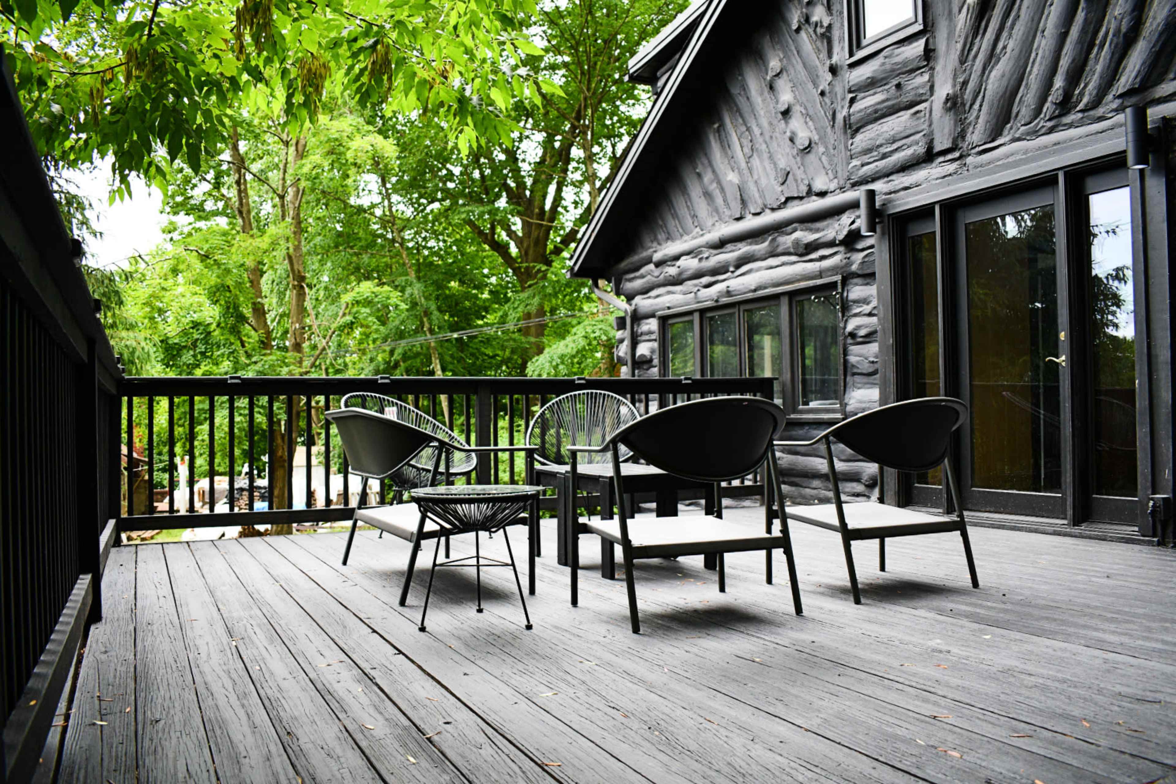 A wooden deck features four modern black chairs and a table, surrounded by lush greenery and a log cabin-style house.