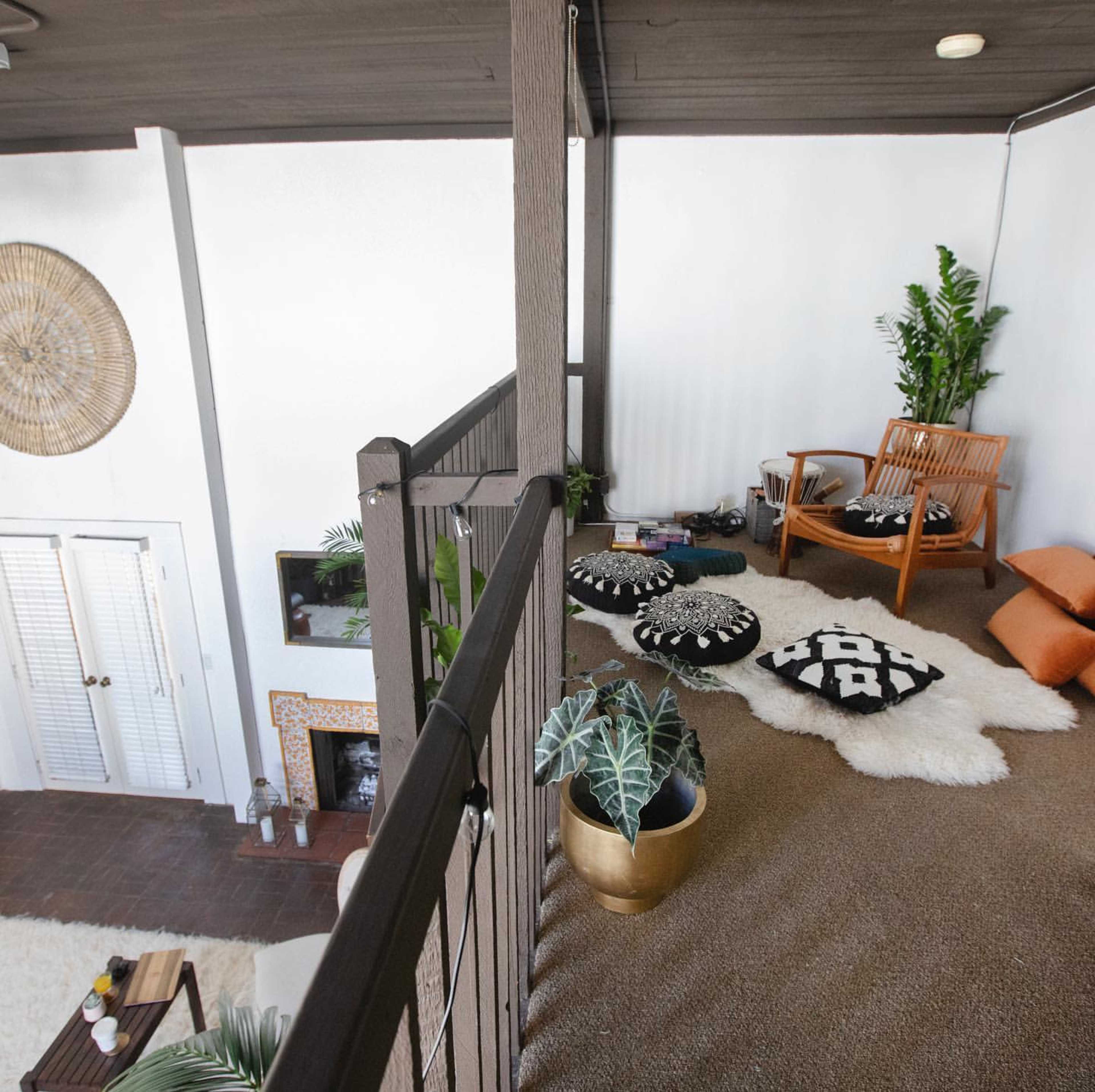 A cozy loft space with a seating area featuring a wooden chair, decorative pillows, a large rug, and several plants.