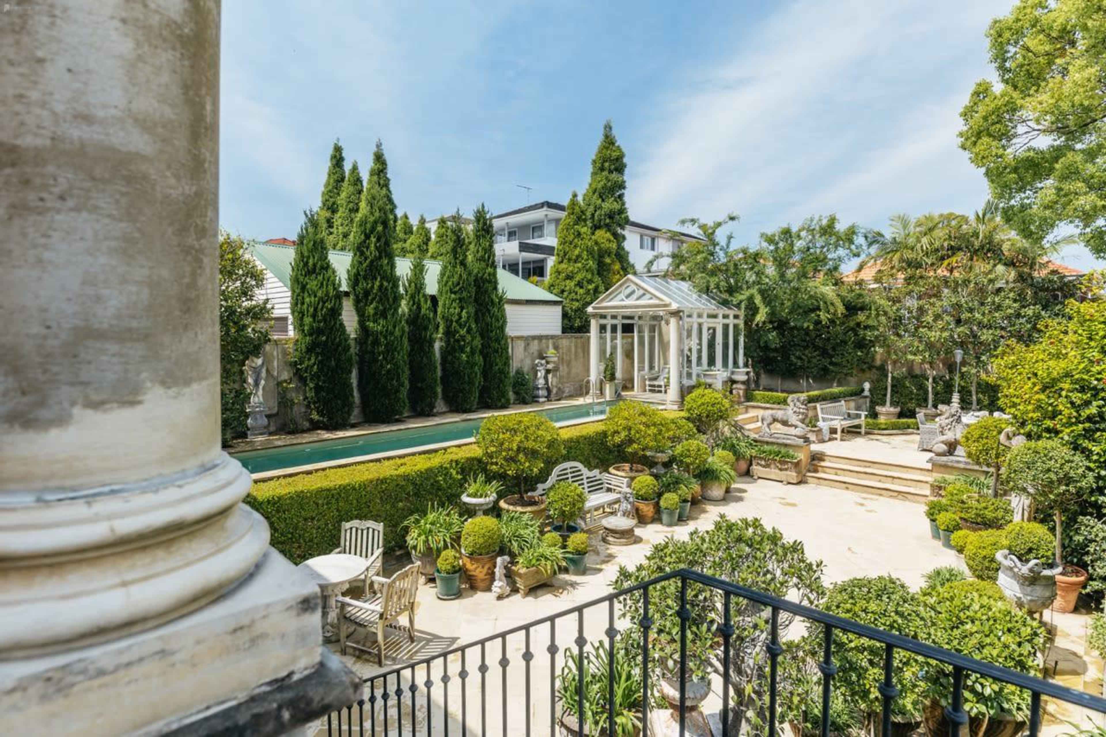 A landscaped garden featuring a long pool, multiple potted plants, and classical sculptures, all surrounded by tall trees and a glass-enclosed structure.