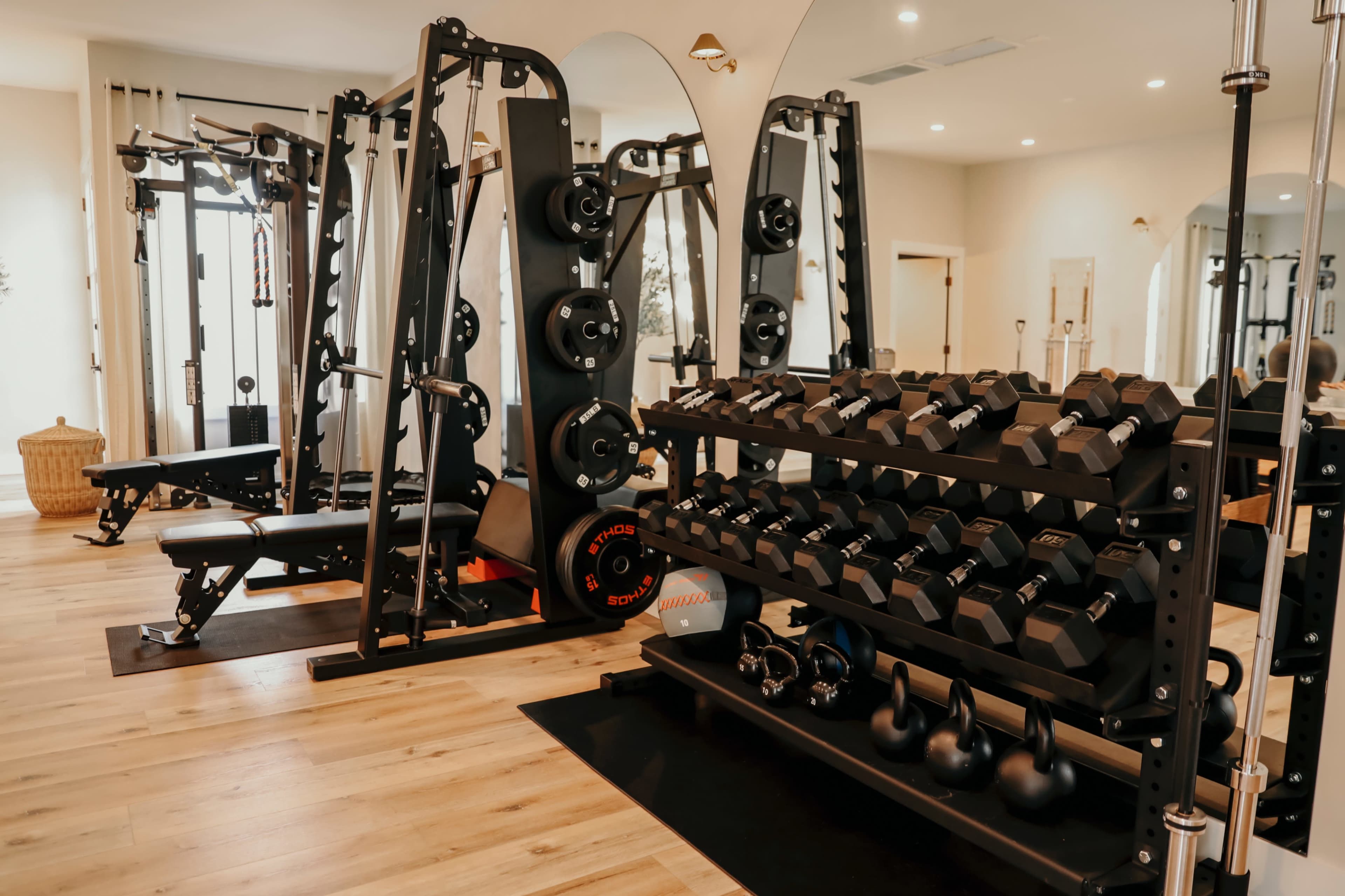 The image shows a well-equipped home gym featuring a weight rack with dumbbells, a multi-functional weight machine, and various workout equipment arranged in a bright, spacious room.