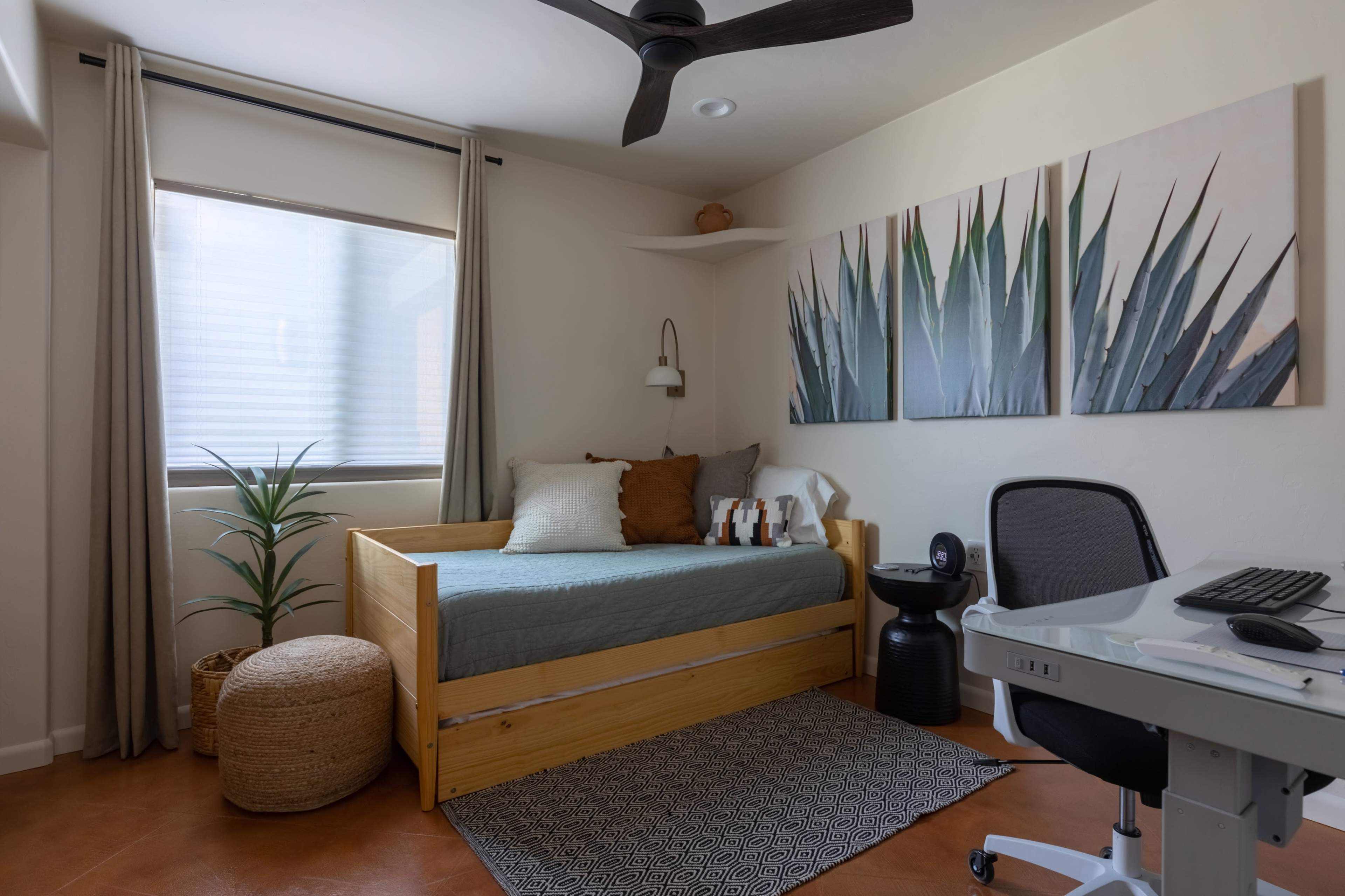 A minimalist bedroom featuring a wooden bed, a small desk with a computer, a plant, and two large wall art pieces depicting greenery.