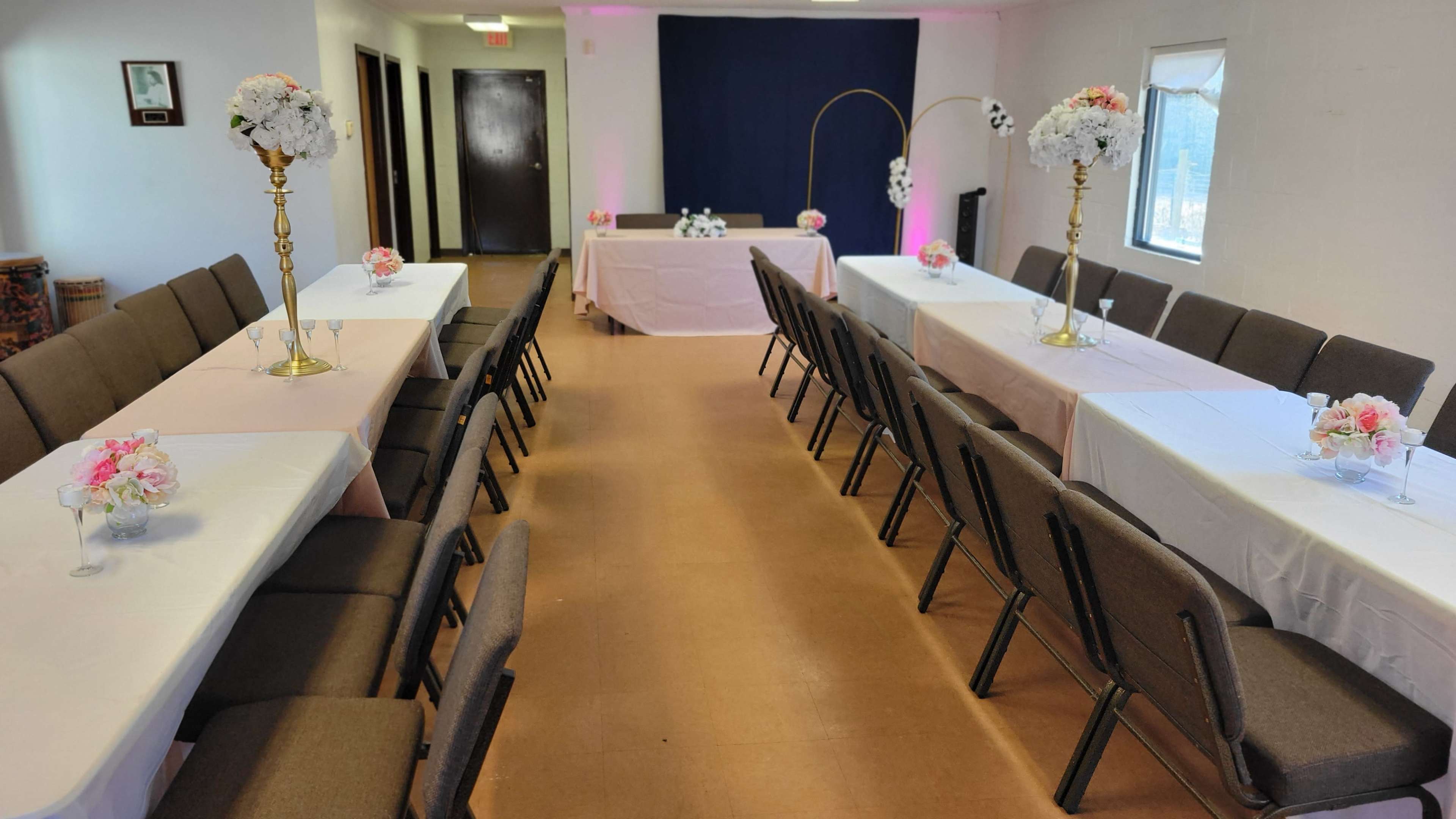 The image shows a decorated indoor event space with two rows of tables set with floral centerpieces and a backdrop for a head table at the front.