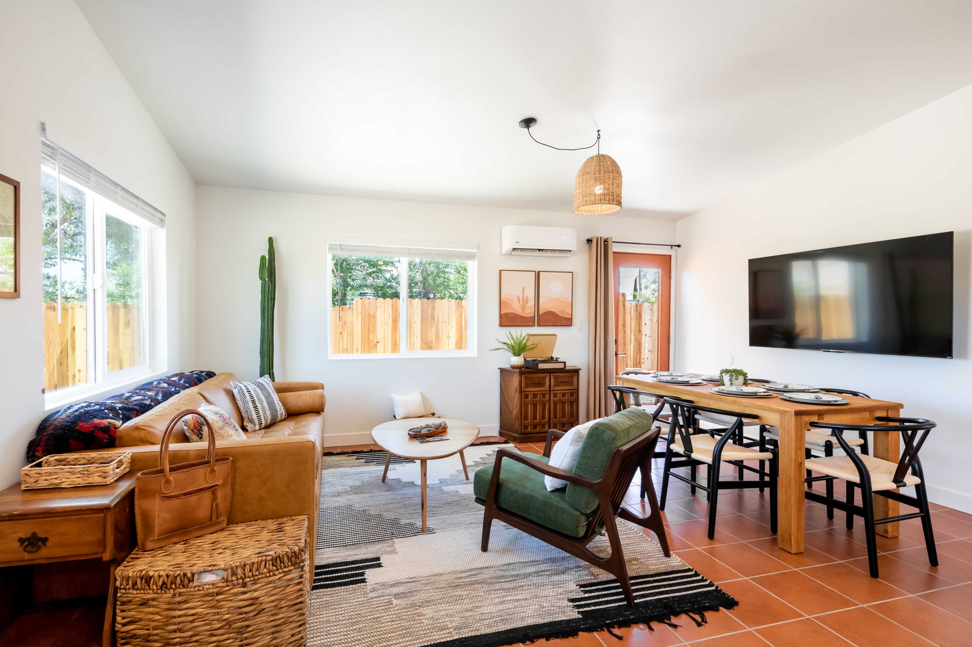 A modern living room features a brown leather sofa, a round coffee table, a dining area with a wooden table, and a large wall-mounted television.